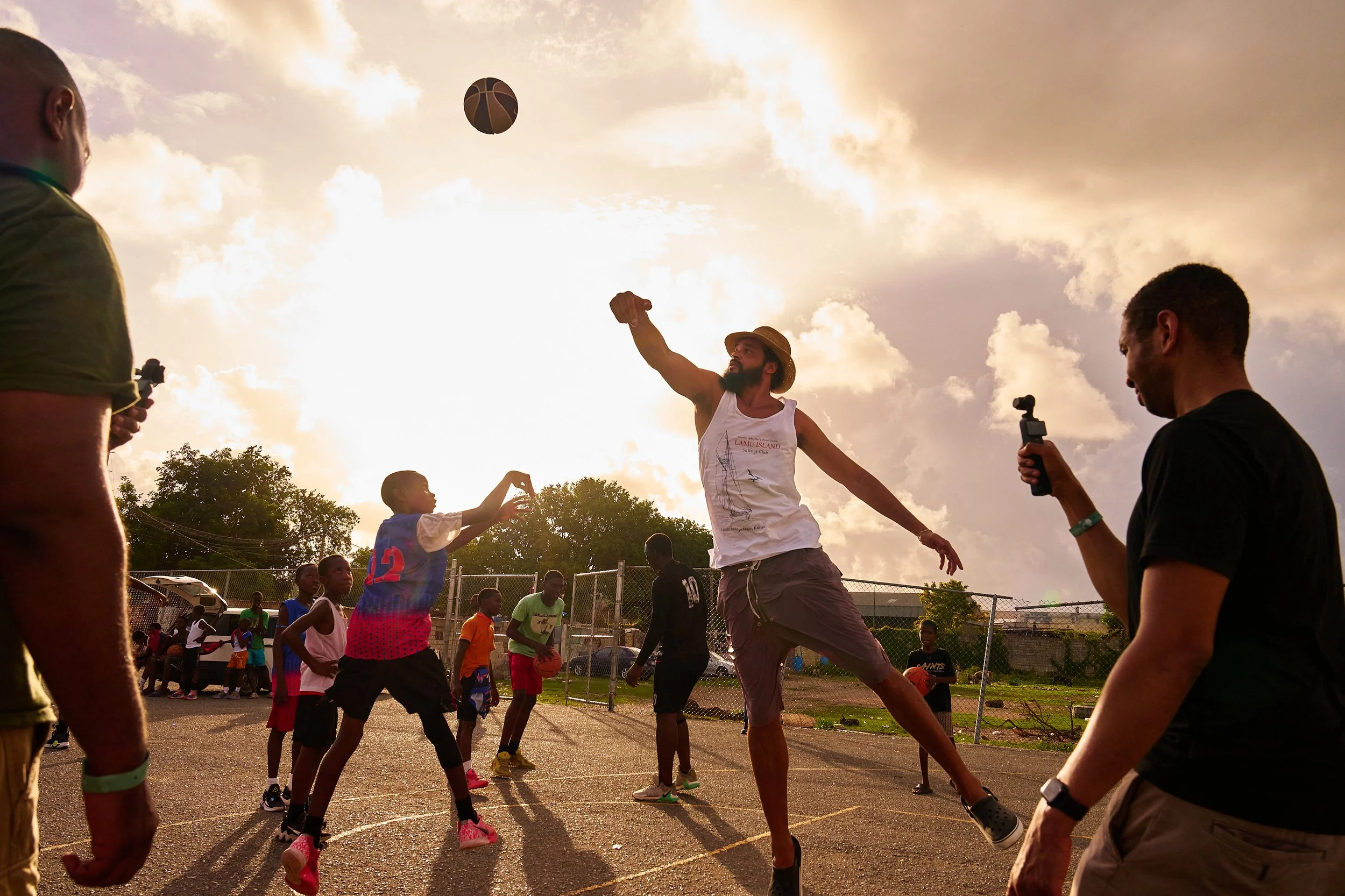 People playing basketball on an outdoor court during sunset, with some players shooting and others watching.