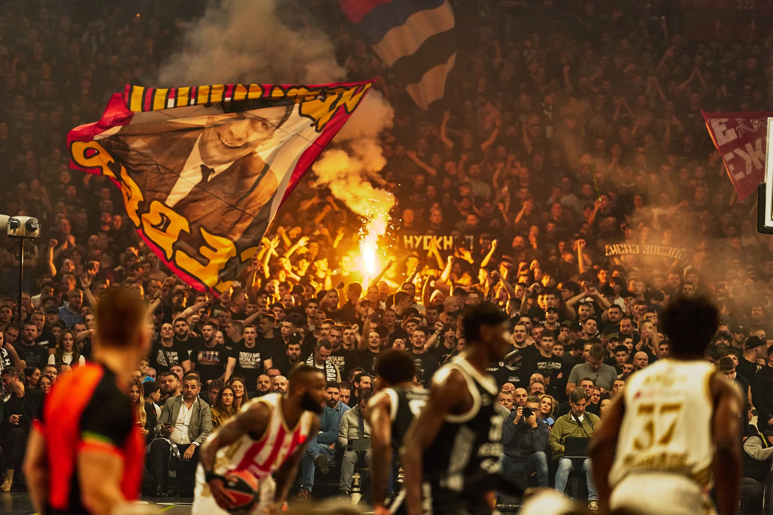 Basketball game with cheering crowd, large flag with a portrait hanging, flares, and teams on the court.