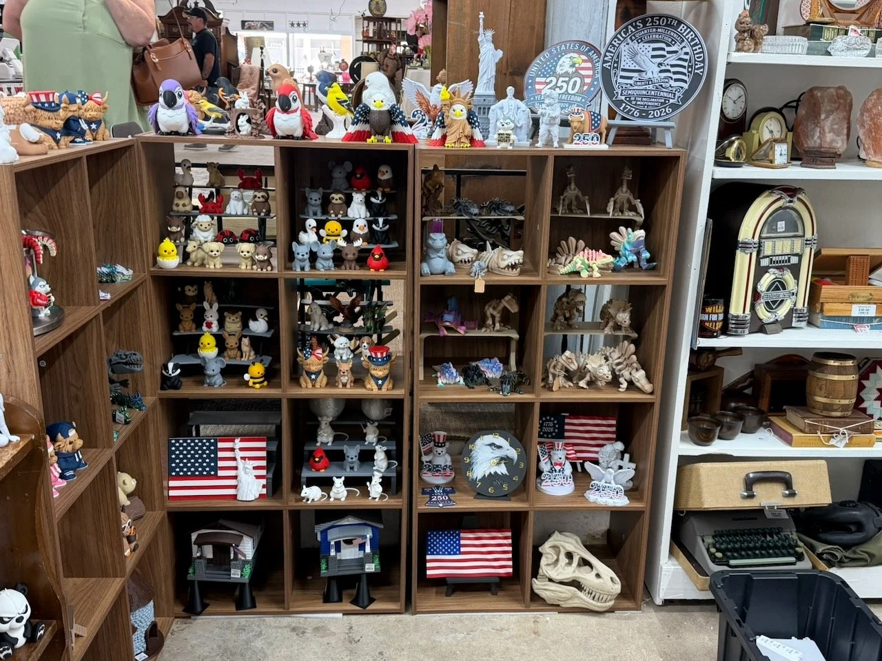 Display of various decorative figurines, collectibles, and clocks on wooden shelves in a store, including plush toys, rock-themed statues, American flags, a clock with an eagle, a vintage jukebox, and a skull replica.