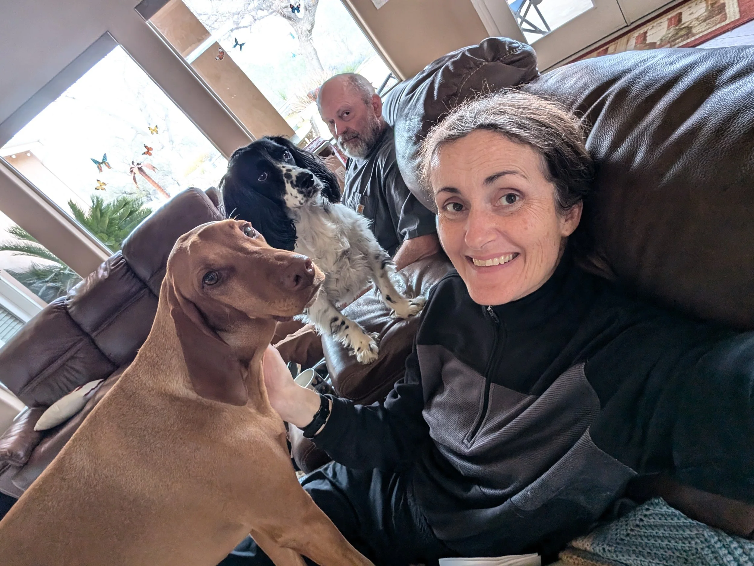 Pet sitter Leona, smiling at the camera with two dogs, and pet sitter Mira, sitting on a couch in a bright living room.