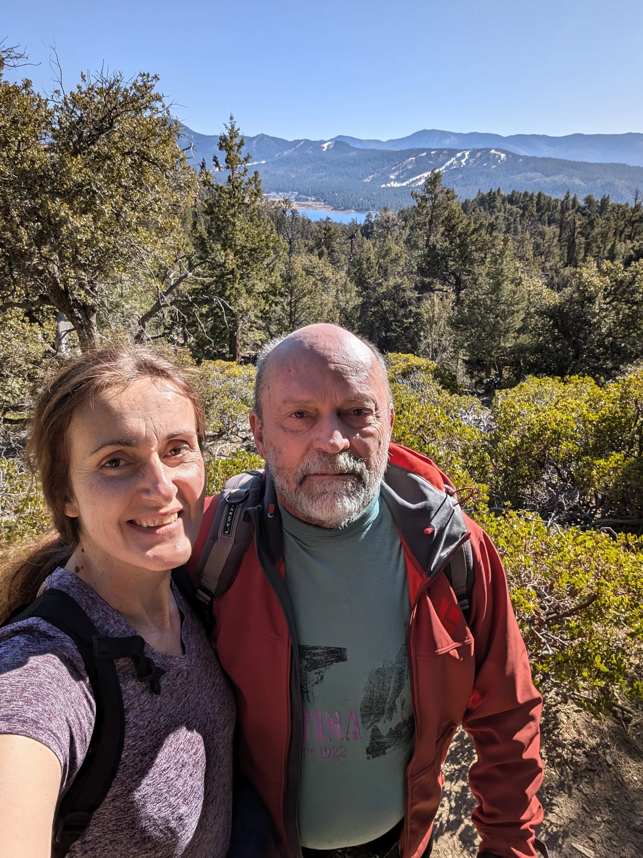Dog walkers, a smiling woman (Leona) and a serious man (Mira), taking a selfie on a hiking trail surrounded by trees and mountains in the background.