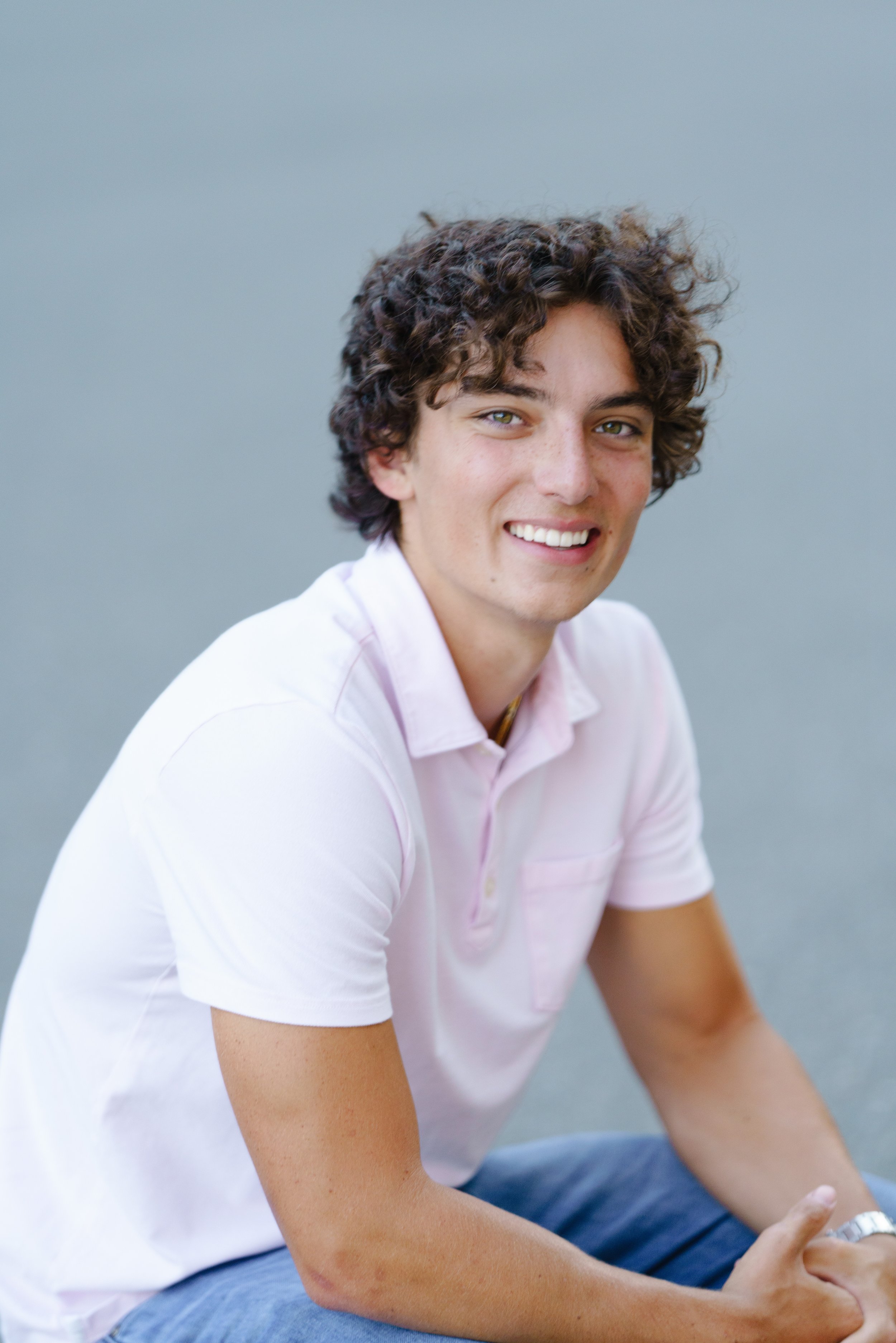 A young man with curly hair and light green eyes smiling while sitting against a plain blue wall, wearing a light pink polo shirt and blue jeans.