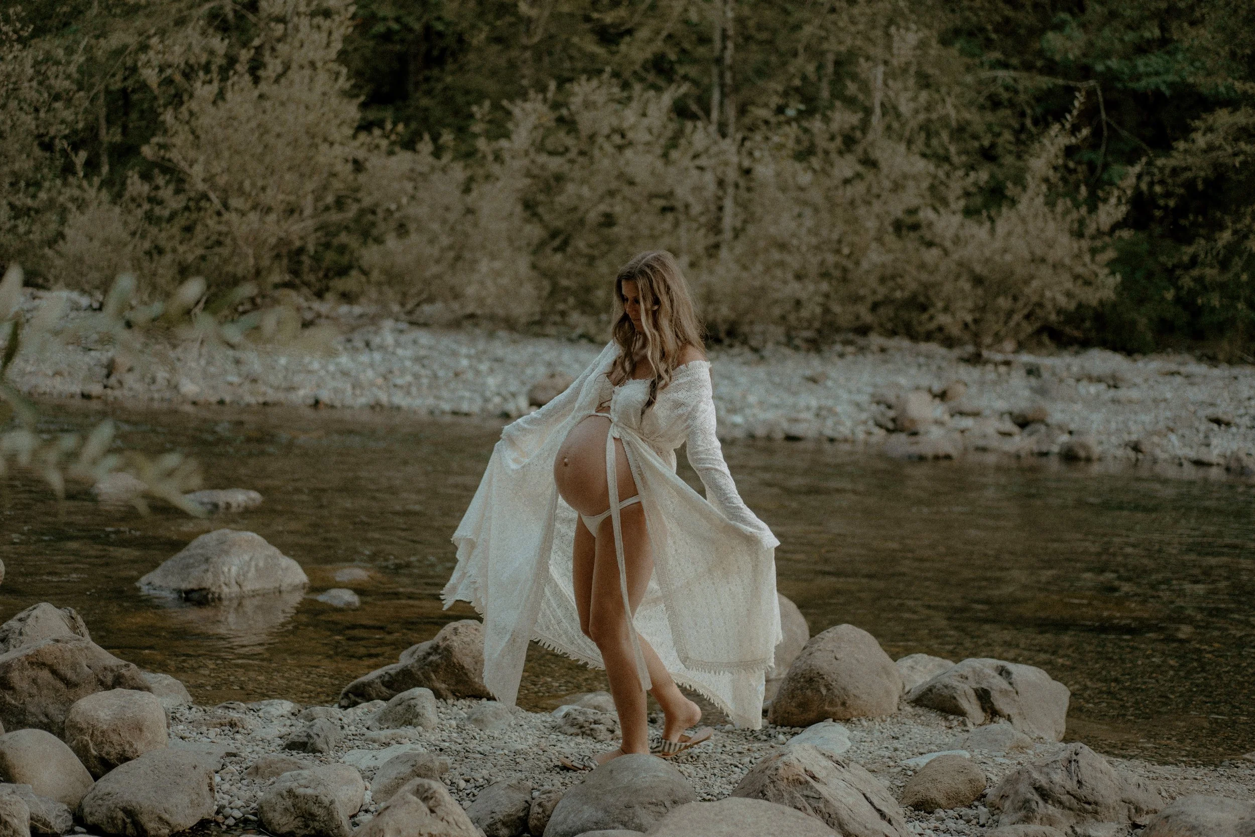 A pregnant woman in a white dress standing on a rocky riverbank with a forest in the background.