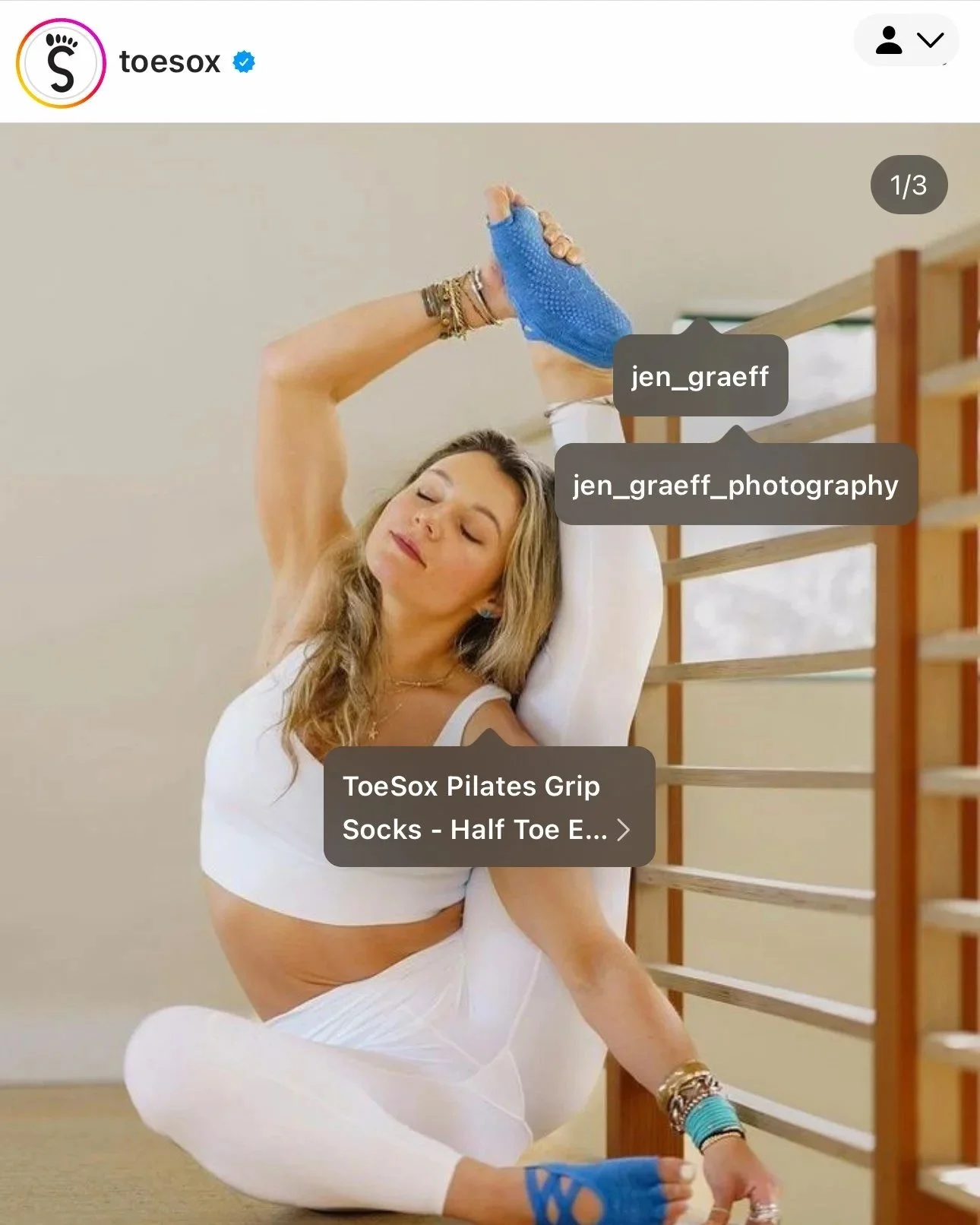 Woman practicing yoga in a white outfit, seated in a twisted pose near a wooden staircase, wearing blue ToeSox Pilates grip socks with half toe design.