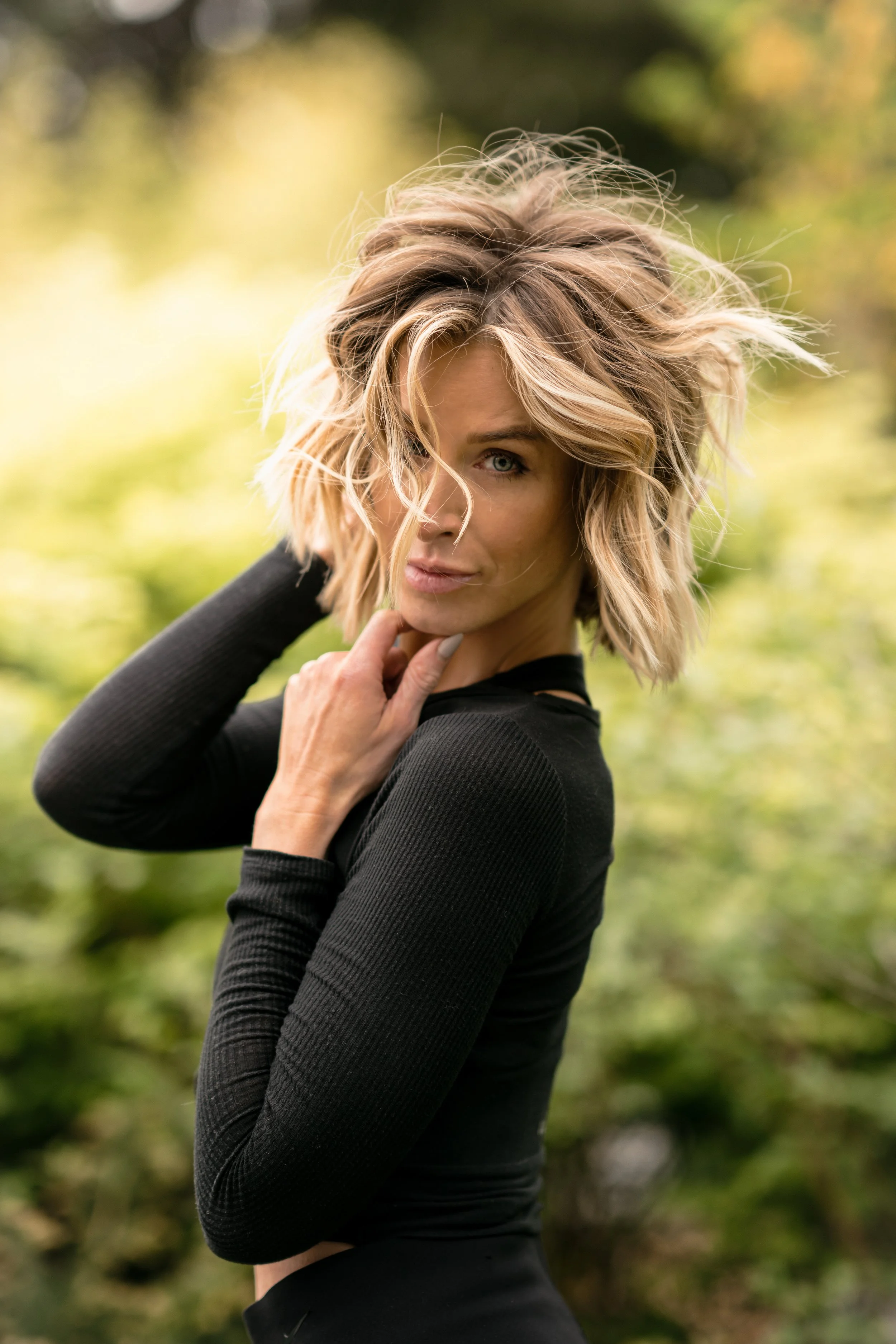 A woman with short, tousled blonde hair wearing a black long-sleeve top is outdoors in a natural setting, with greenery and blurred foliage in the background.