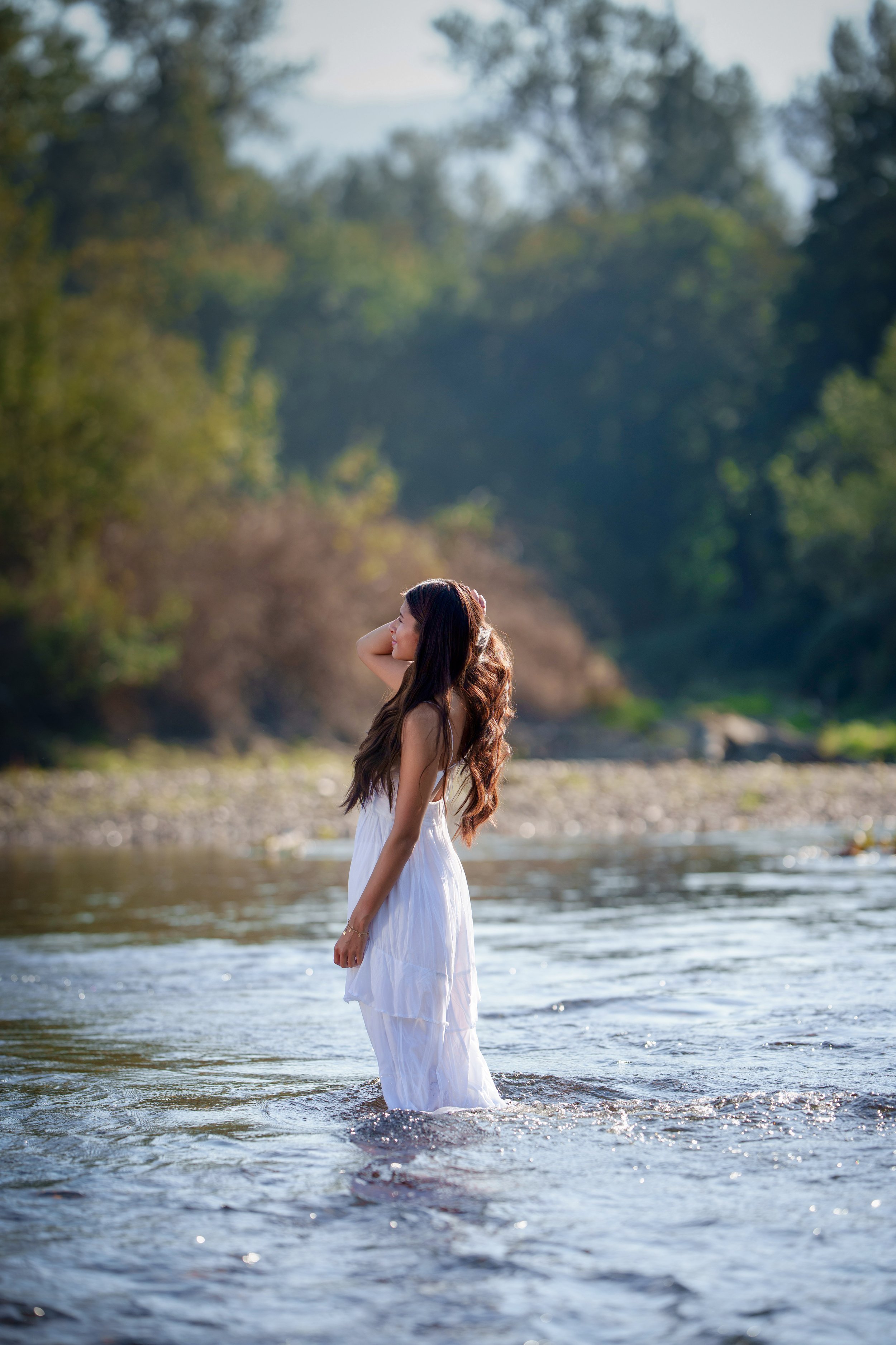 A woman in a white flowing dress standing in a shallow river, with a background of trees and a clear sky.