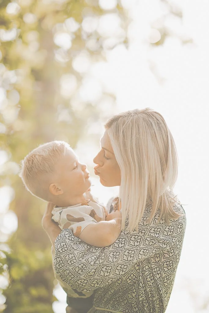 A woman and a young child are outdoors, facing each other closely, with the woman holding the child in her arms. They are about to kiss or touch noses, with a background of blurred trees and sunlight filtering through.
