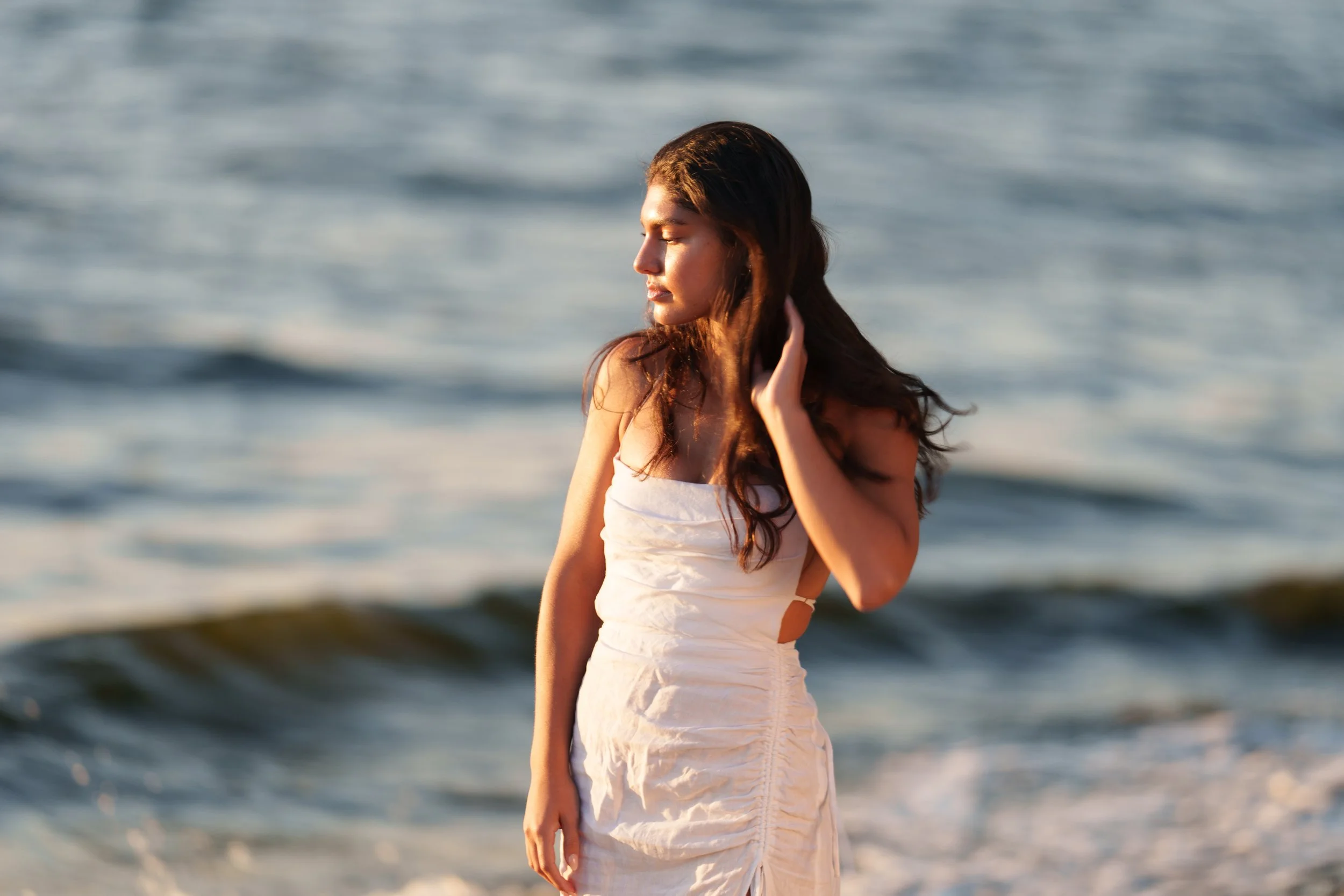 A woman in a white dress with chestnut hair standing at the beach with the ocean waves in the background, her eyes closed and touching her hair.