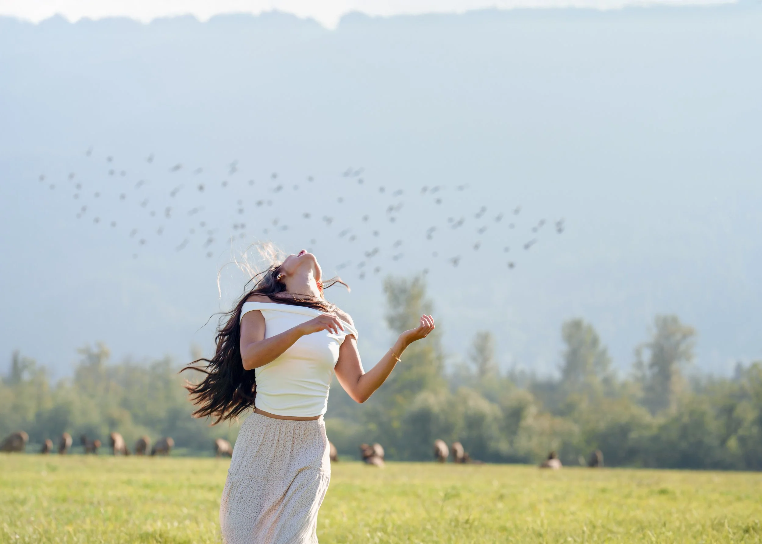 A woman with long hair wearing a white top and a beige skirt is standing outdoors in a field during daytime, with her head tilted back and eyes closed, enjoying the moment. In the background, there is a herd of cattle grazing, and a flock of birds flying in the sky.