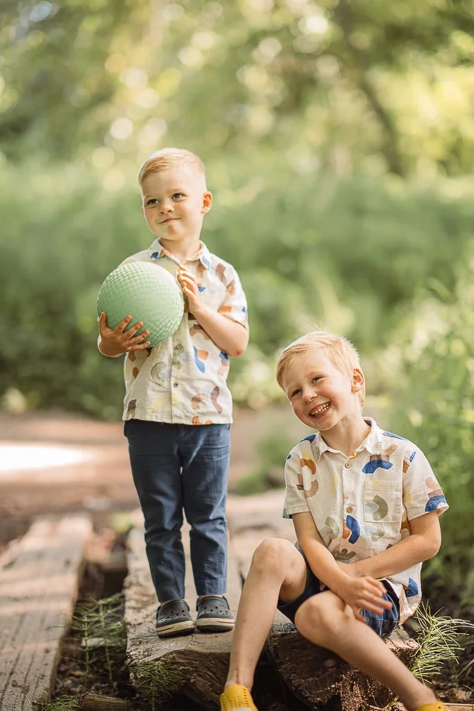 Two young boys with blond hair and light skin in patterned shirts outdoors on a boardwalk surrounded by greenery. One boy is standing, holding a green ball, and the other is sitting on a log, smiling.