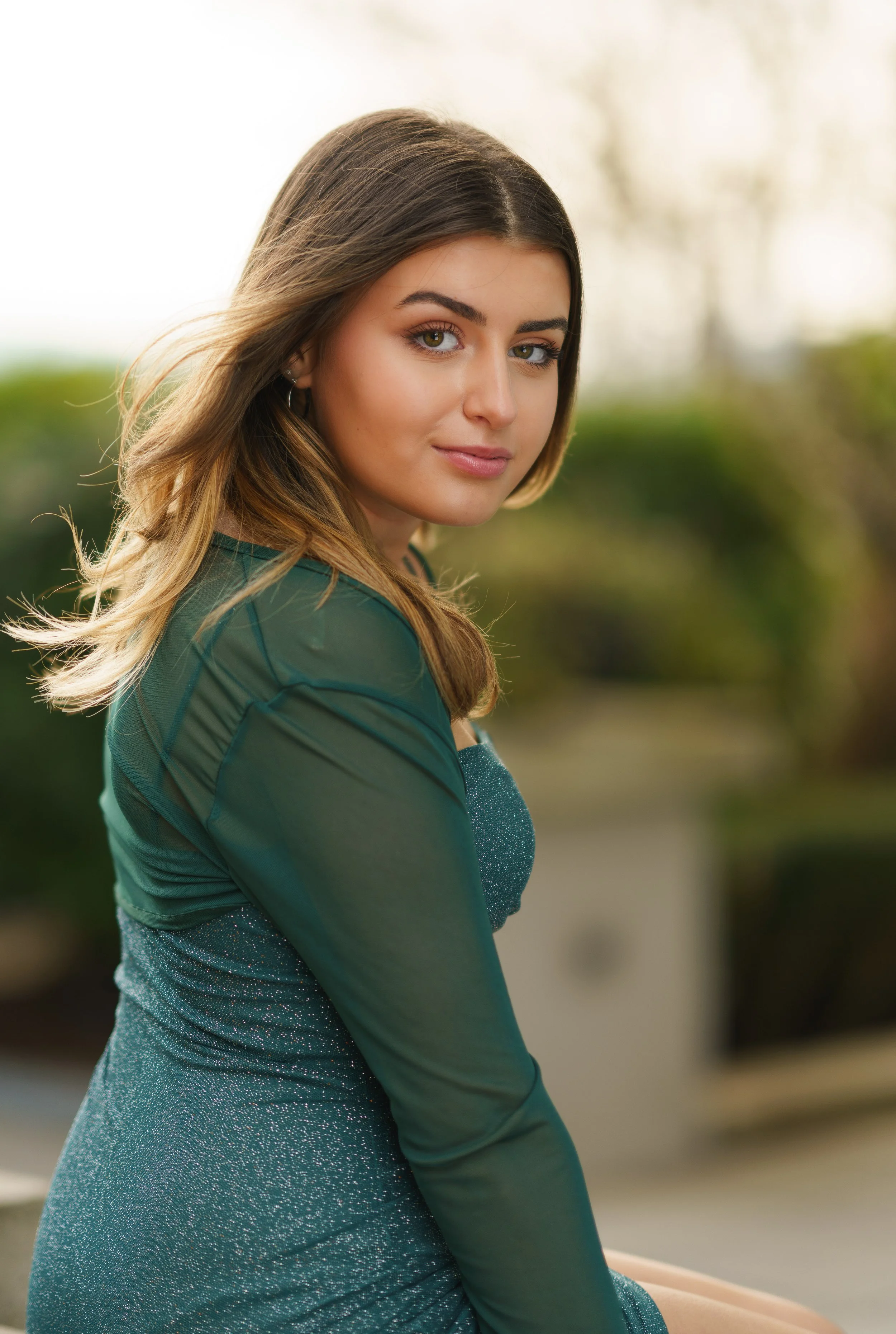 A young woman with long, brown hair and hazel eyes wears a green top and a sparkly blue dress, standing outdoors with a blurred background of trees and greenery.