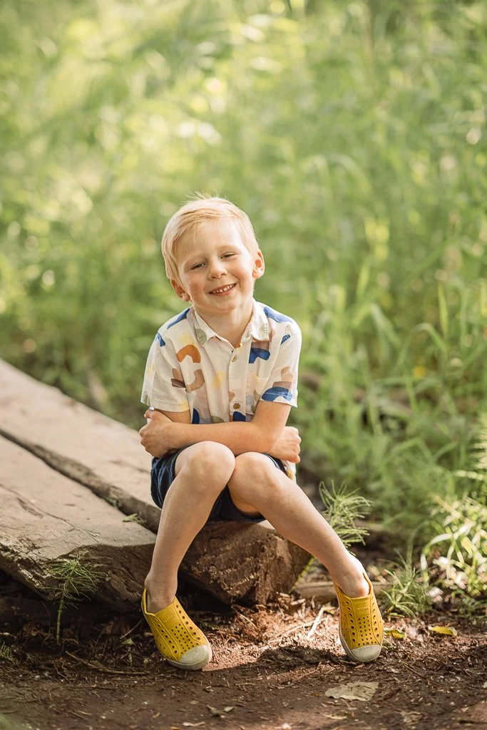 A young boy sitting on a log outdoors, smiling at the camera, surrounded by trees and greenery.