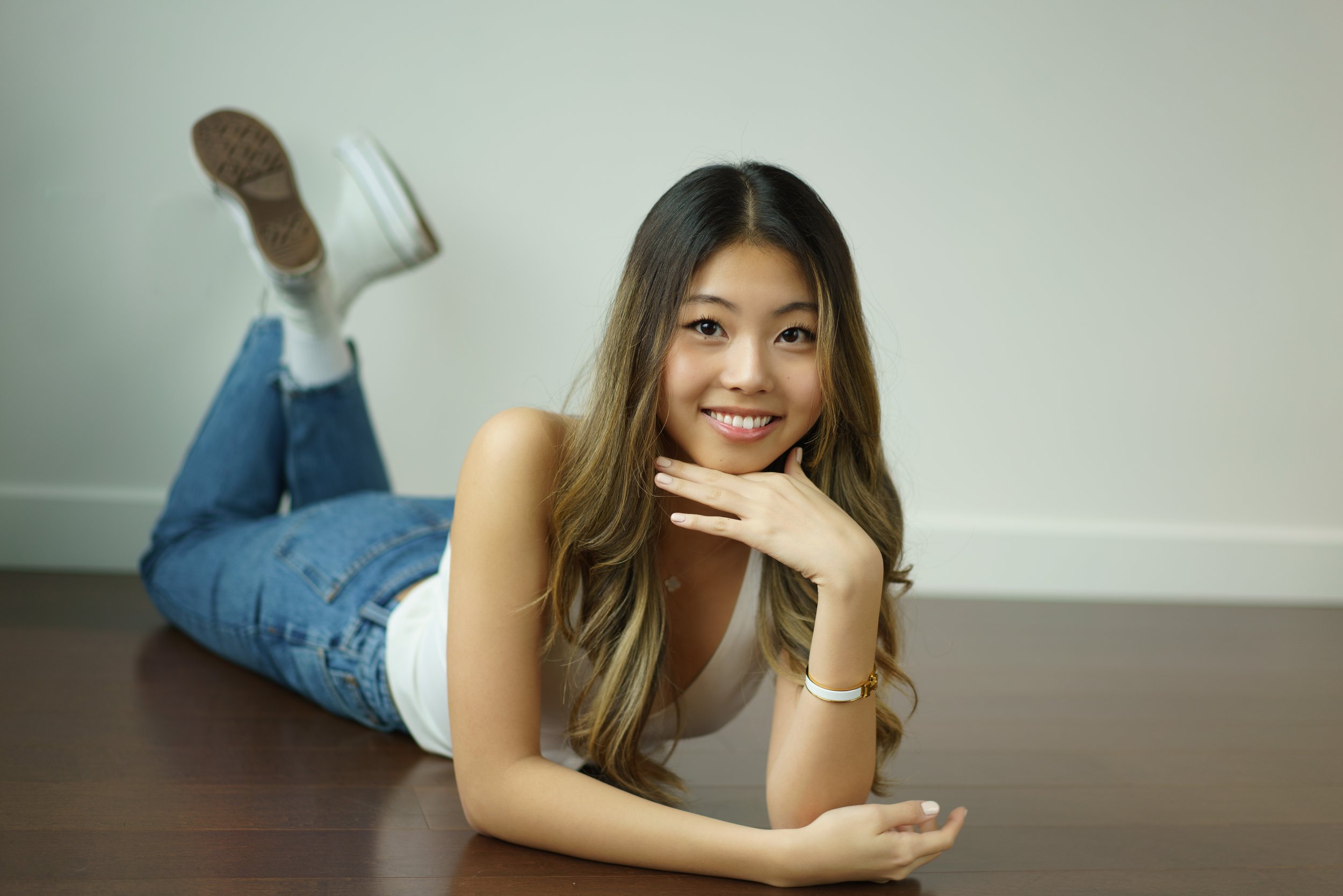 Young woman lying on her stomach on a dark wooden table, smiling at the camera, with her head resting on her hand, wearing a white tank top, blue jeans, and white sneakers.