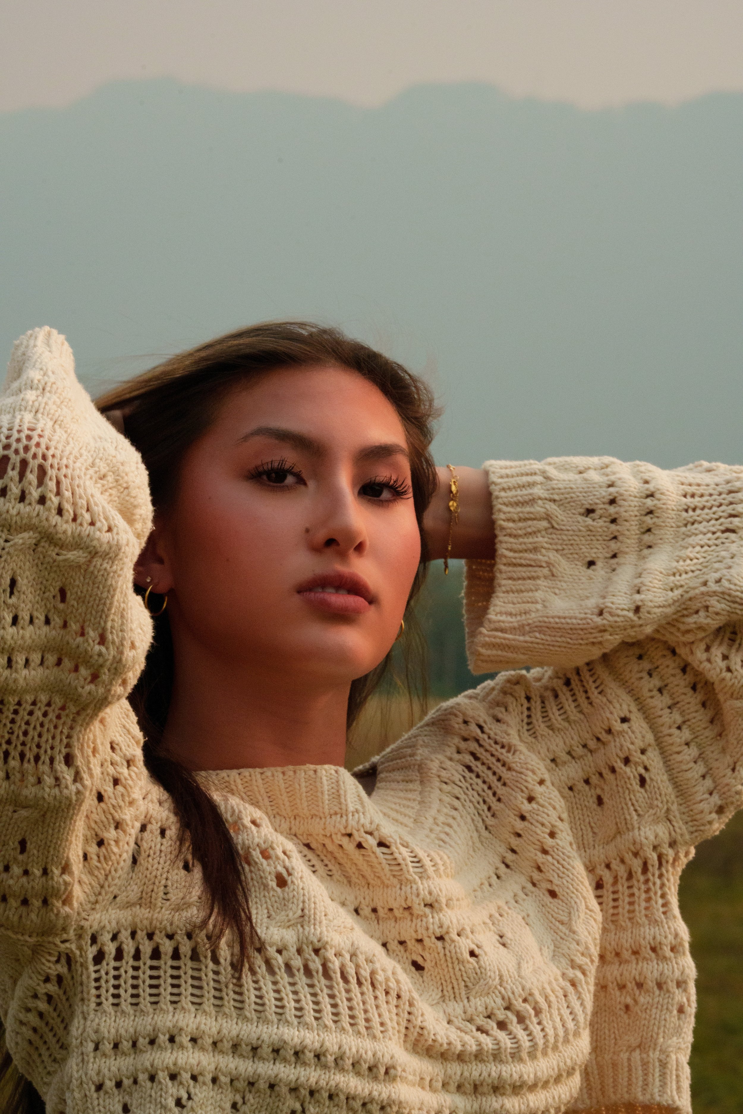 A young woman with long dark hair and makeup, wearing a cream-colored knitted sweater, posing outdoors with her arms raised behind her head, against a soft-focus natural background during sunset.
