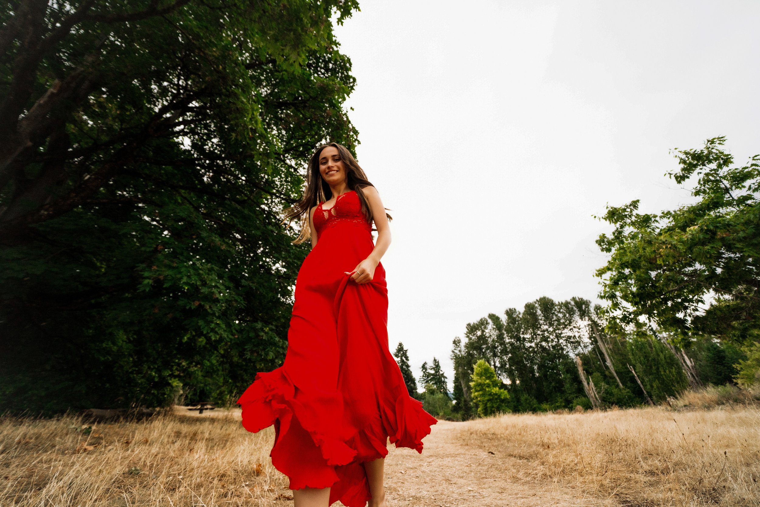 A woman wearing a long red dress standing outdoors in a natural setting, with trees and a cloudy sky in the background.