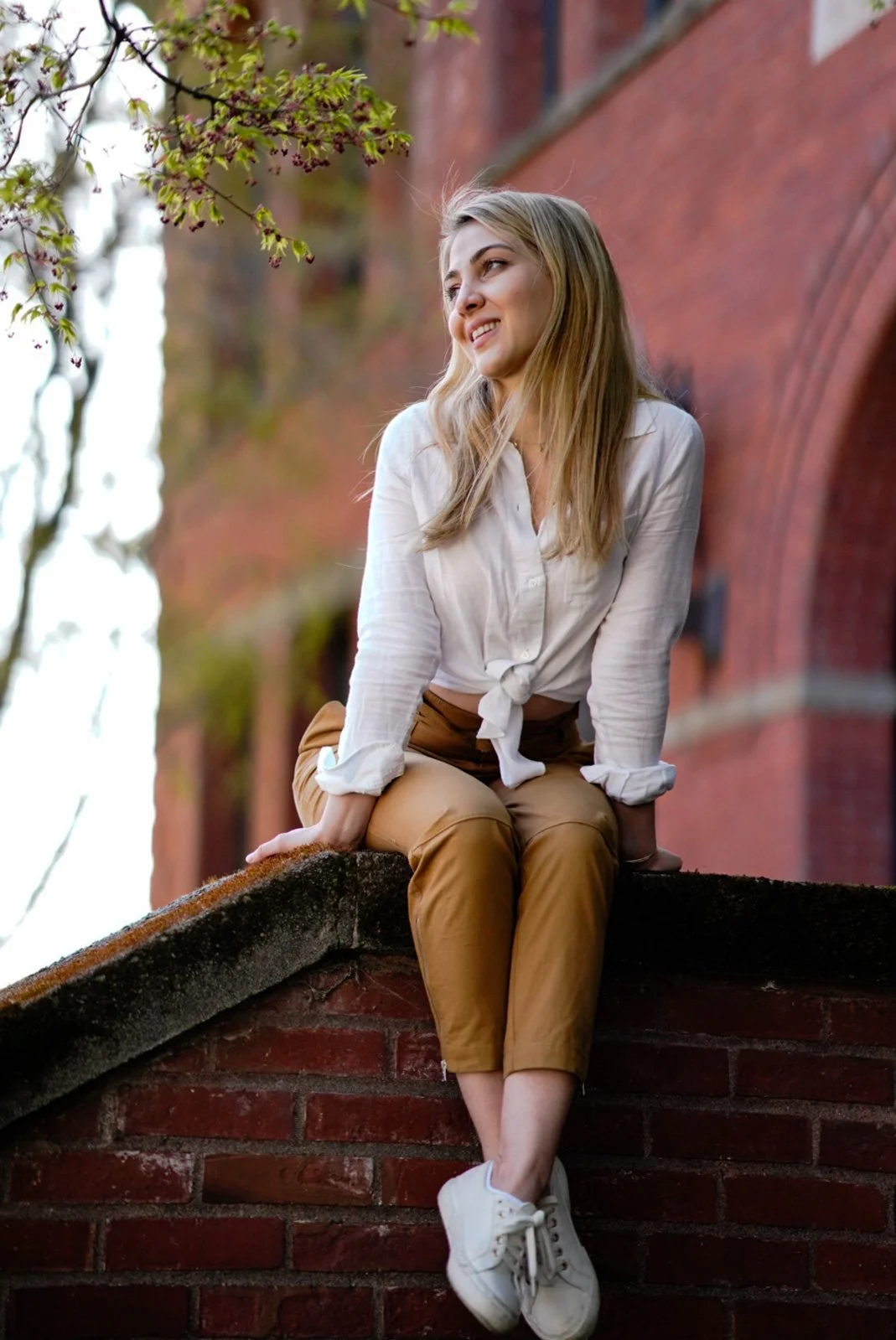 A young woman with long blonde hair casually dressed in a tied white blouse, tan pants, and white sneakers, sitting on a brick ledge with a red brick wall and tree with green leaves in the background.