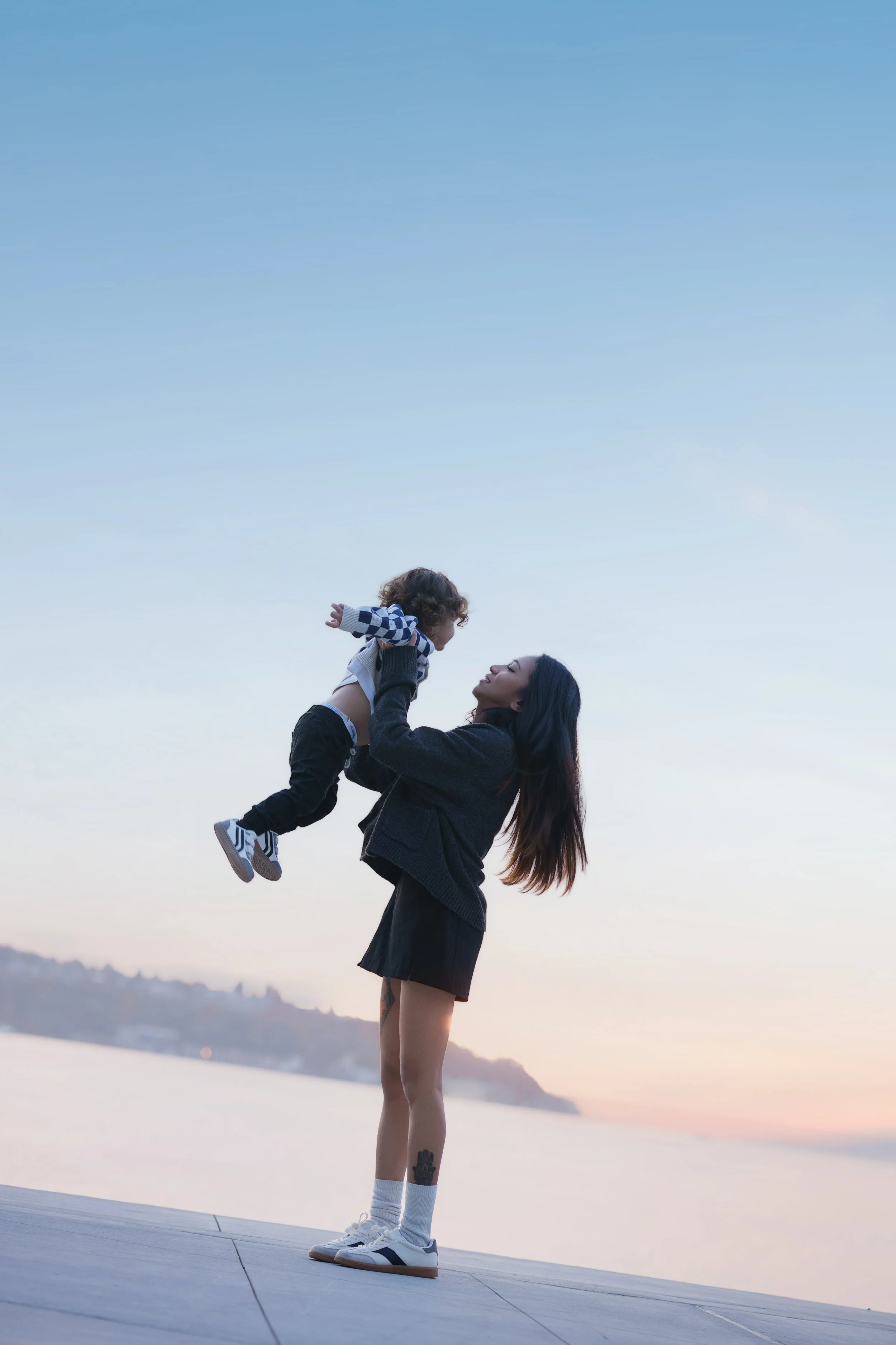 A woman holding a young child in the air while standing outdoors on a wooden path, with mountains and water in the background during sunset.