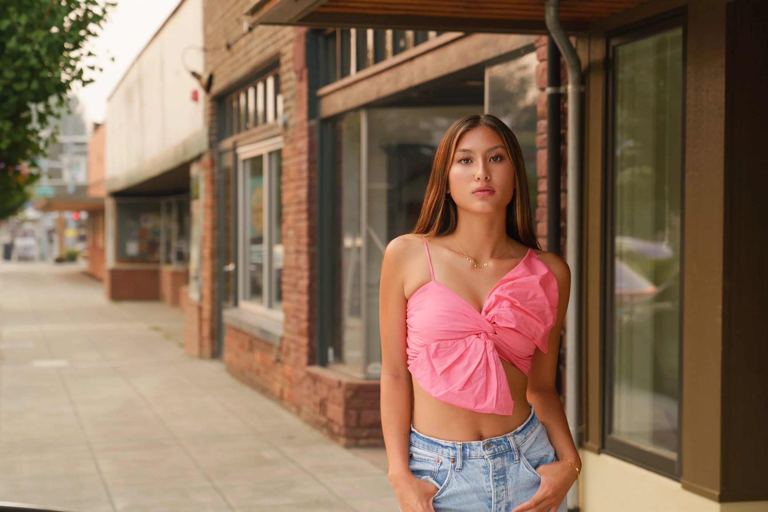 A young woman standing on a sidewalk in front of a brick building with large windows, wearing a pink top and jeans, with her hands in her pockets, looking at the camera.