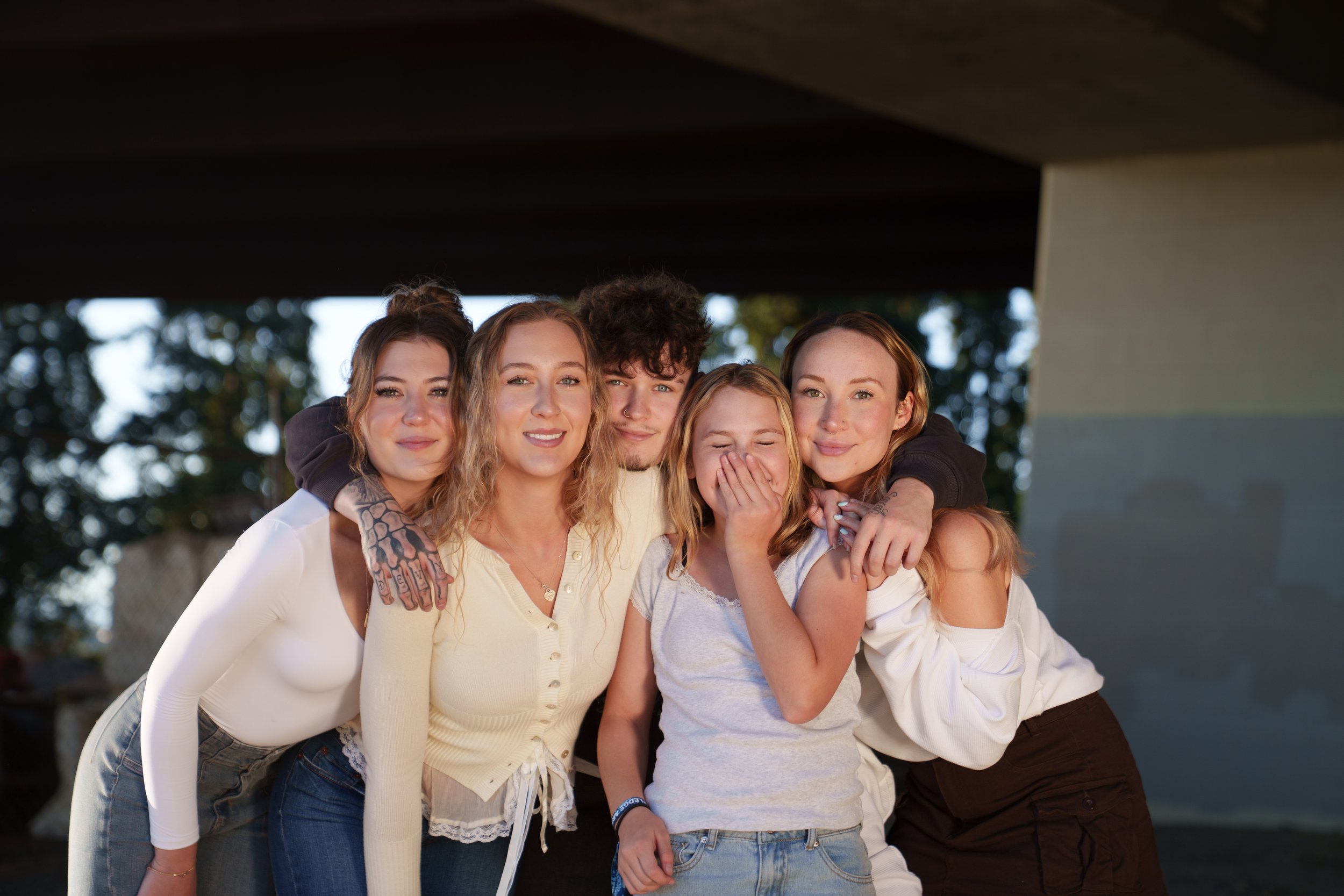 A group of five diverse young friends, smiling and hugging outdoors under a bridge with trees in the background.