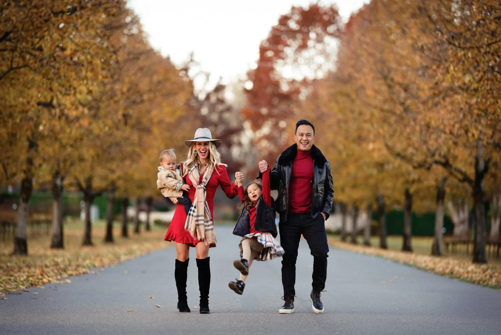 A family of four walking and playing outdoors on an autumn day, with trees with colorful fall leaves in the background. The woman is holding a toddler, the girl is swinging between the woman and man, and they are all smiling.