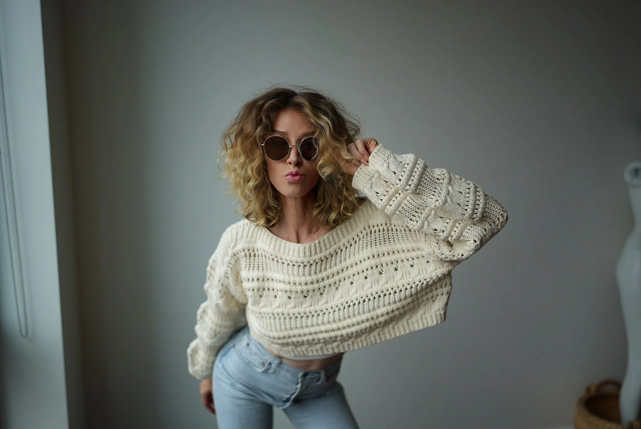 A woman with curly blonde hair wearing dark sunglasses, a cream knitted sweater, and light blue jeans posing with one hand on her sunglasses and lips pursed, standing indoors against a gray wall.