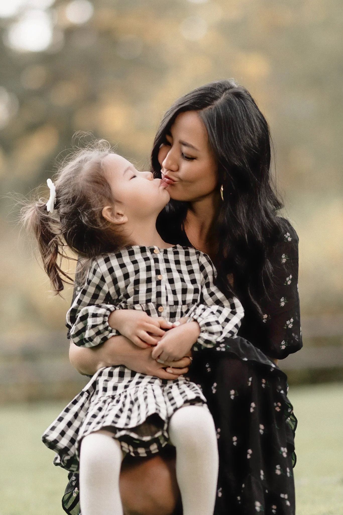 A woman with long black hair holding a young girl with light brown hair in ponytails, wearing a checkered dress, outdoors against a background of blurred trees and natural scenery, sharing a kiss.