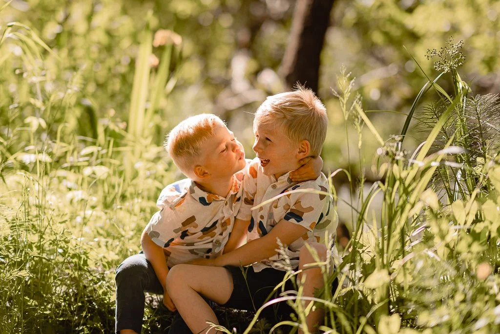 Two young boys with blonde hair, wearing matching camo shirts, sitting on the grass in a sunny, green outdoor setting, playfully fighting or teasing each other.