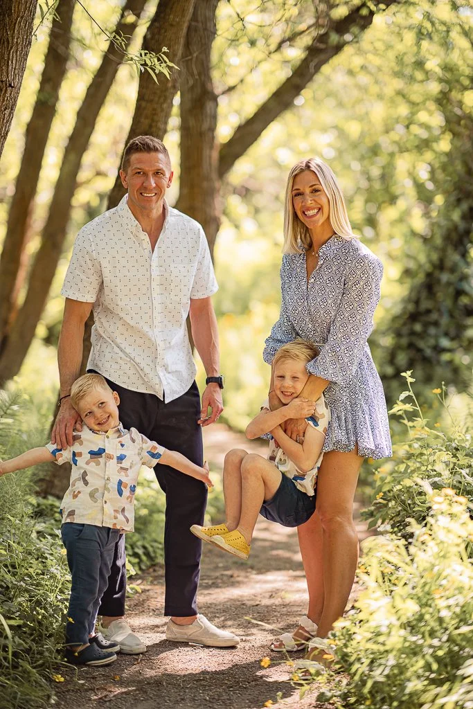 A happy family of four, including two young children, poses on a sunlit forest path surrounded by trees and greenery. The parents are smiling, with the mother holding a girl and the father standing beside a boy.