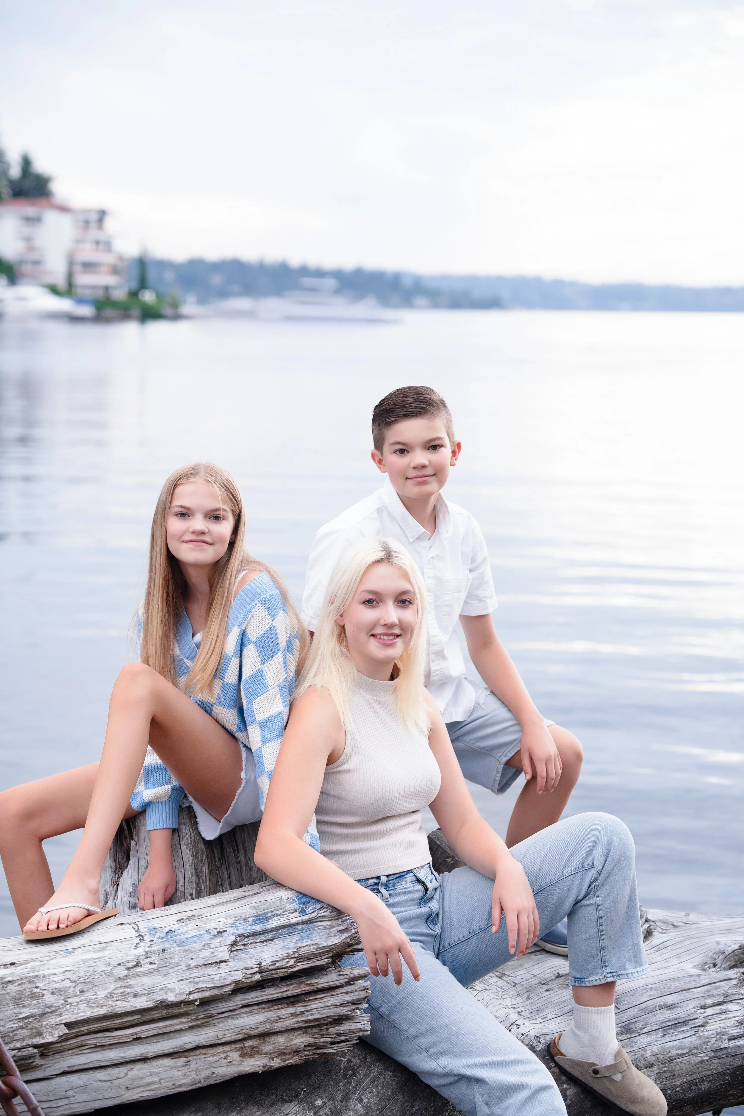 Four young people sitting on a weathered log by a body of water with buildings and trees in the background during daytime.