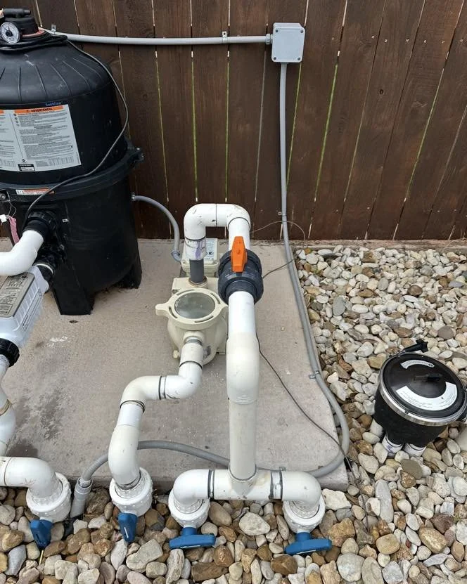 Pool pump system with various pipes, valves, and a filter on a concrete pad beside a wooden fence, surrounded by rocks.