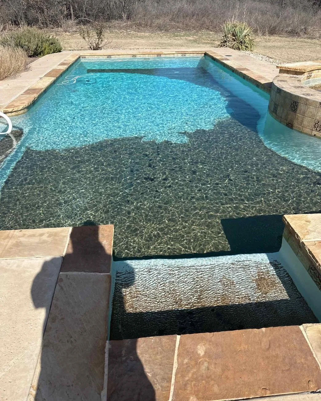 An outdoor swimming pool with a shallow area featuring a textured black and brown surface, and a deeper blue area. The pool is surrounded by a stone patio, with dry grass and shrubs in the background under clear weather.