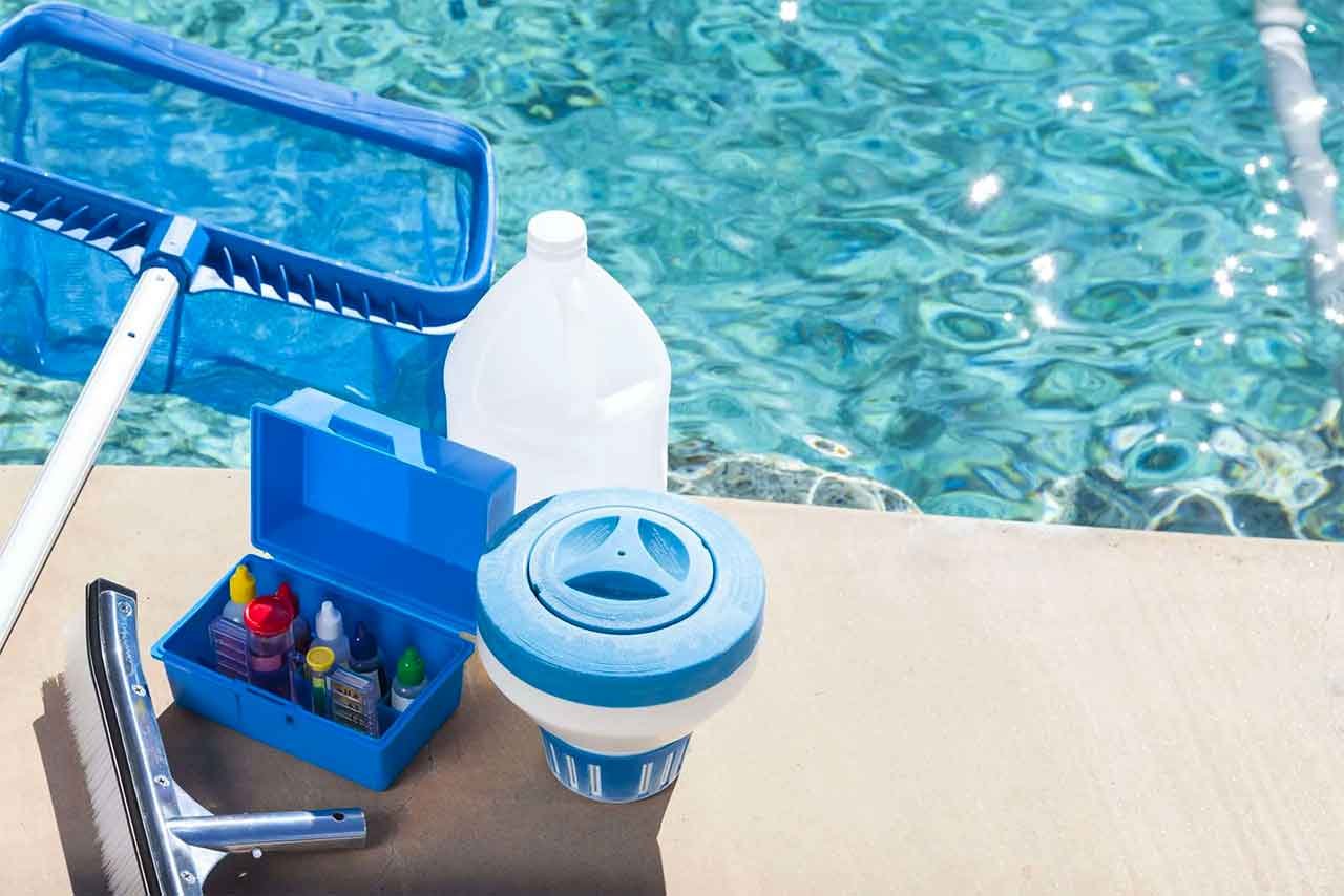 Pool cleaning supplies on a pool deck, including a pool skimmer net, a bottle of pool chemicals, a container with various chemicals, and a pool filter or pump component, with the swimming pool water in the background.