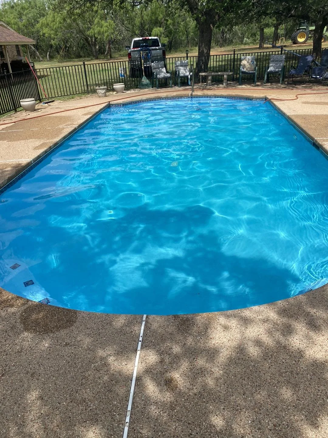 In-ground rectangular swimming pool with blue water, surrounded by a concrete deck, black metal fence, and outdoor furniture, with trees and a tractor in the background.
