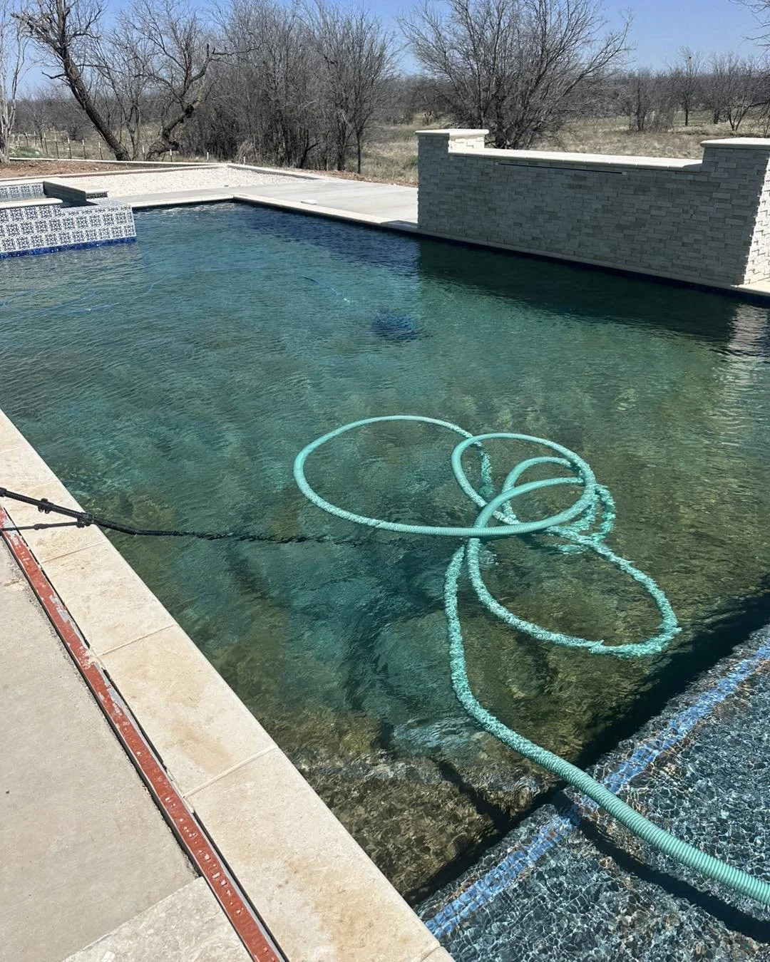 A backyard swimming pool with a clear view of the water, a pool cleaning hose submerged, and a stone wall in the background, set in a rural area with leafless trees.