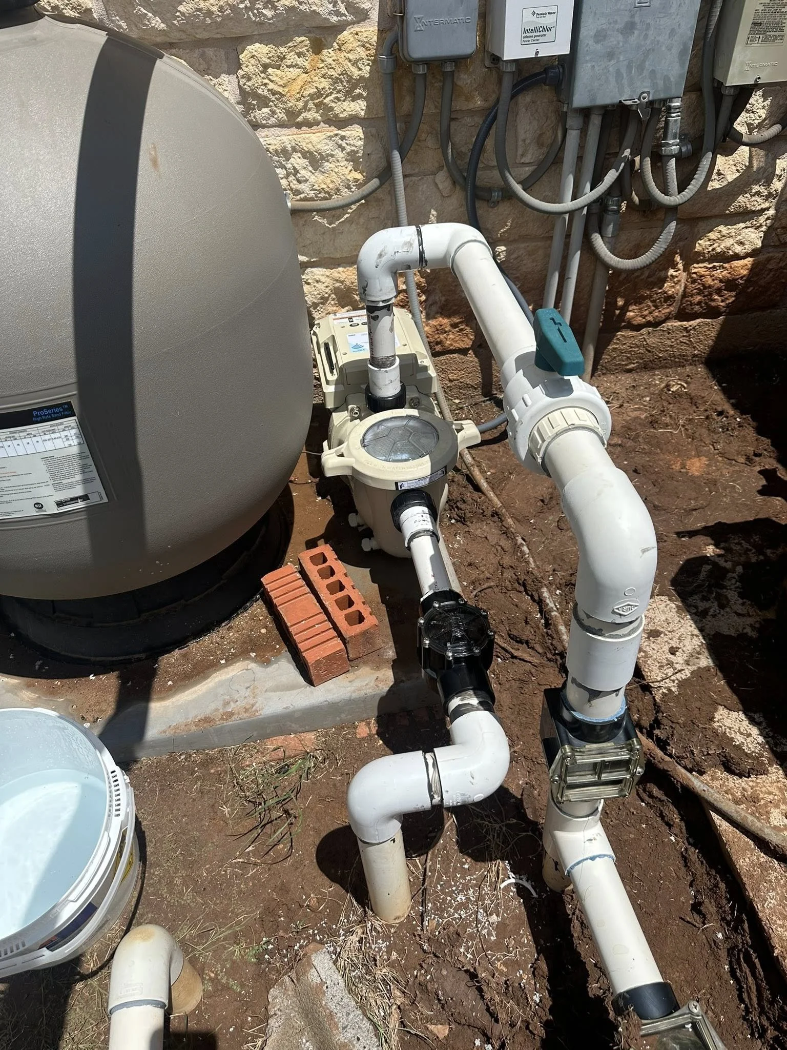 Pool equipment, including a pool pump and filter, with PVC pipes, electrical boxes, and wiring on a dirt ground outside a stone wall.