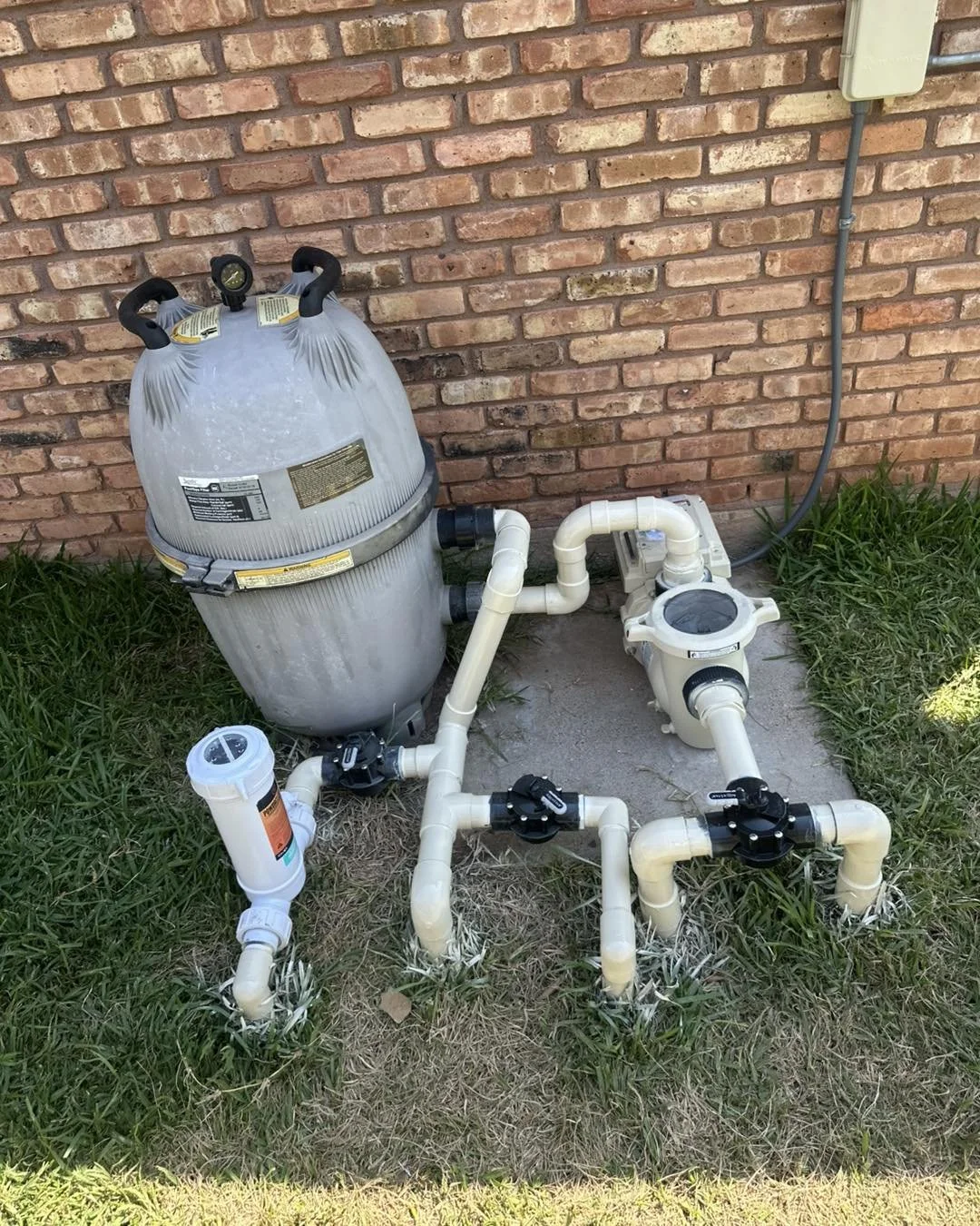 Above ground swimming pool pump and filter system with piping installed next to a brick wall with grass on the ground.