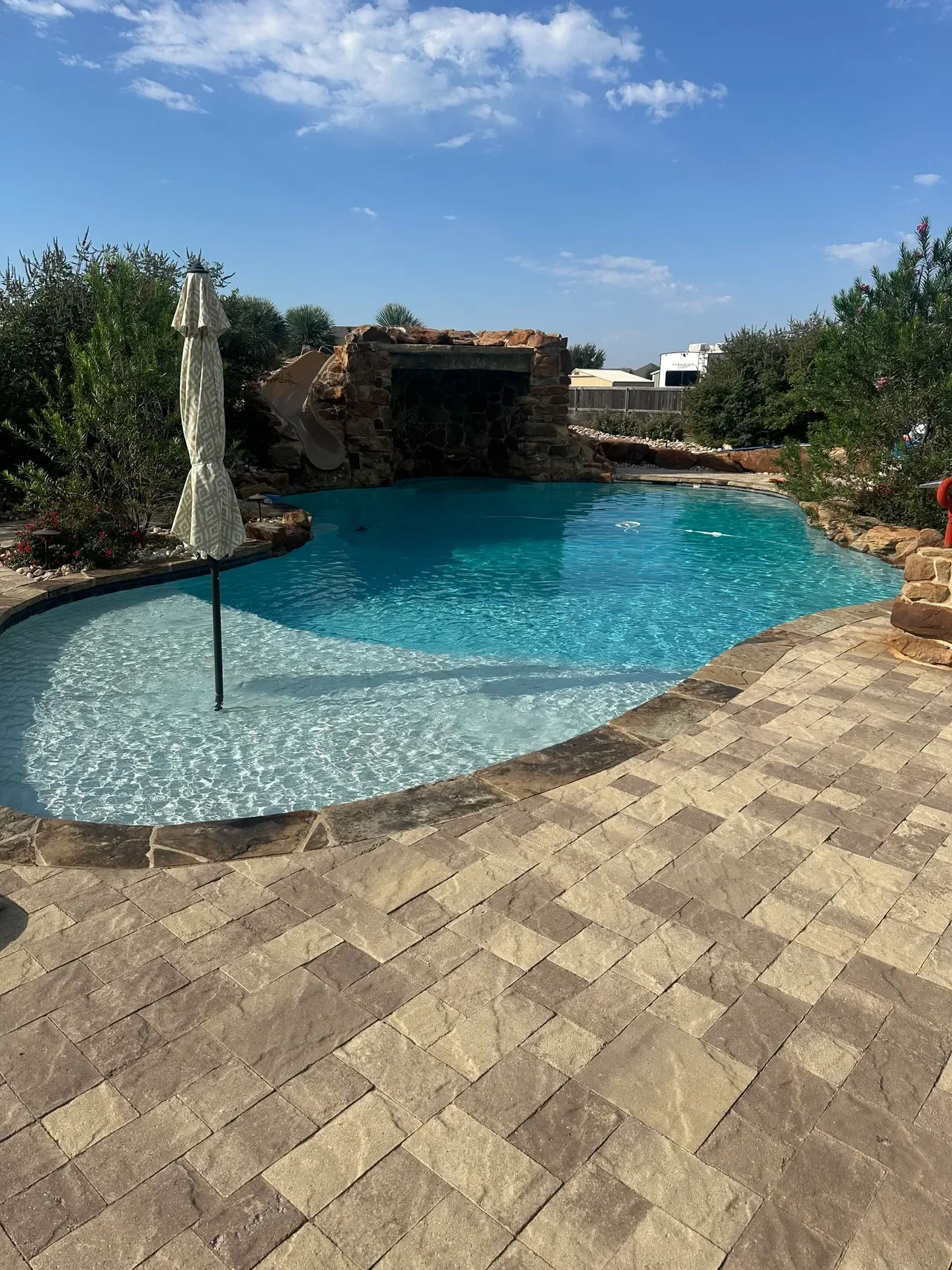 A backyard swimming pool with a shallow entry area, a rock water slide, and a stone water feature. The pool is surrounded by a stone patio with plants and bushes around it. The sky is clear with a few clouds.