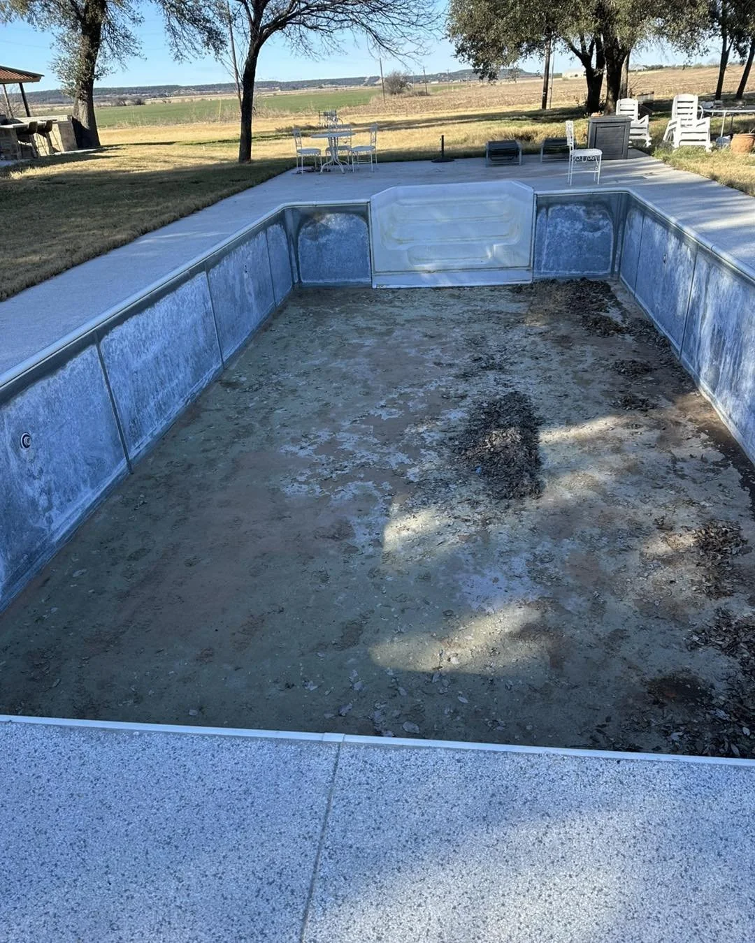 Empty swimming pool with peeling paint and dirt on the bottom, surrounded by a concrete deck, with trees, chairs, and a table in the background.