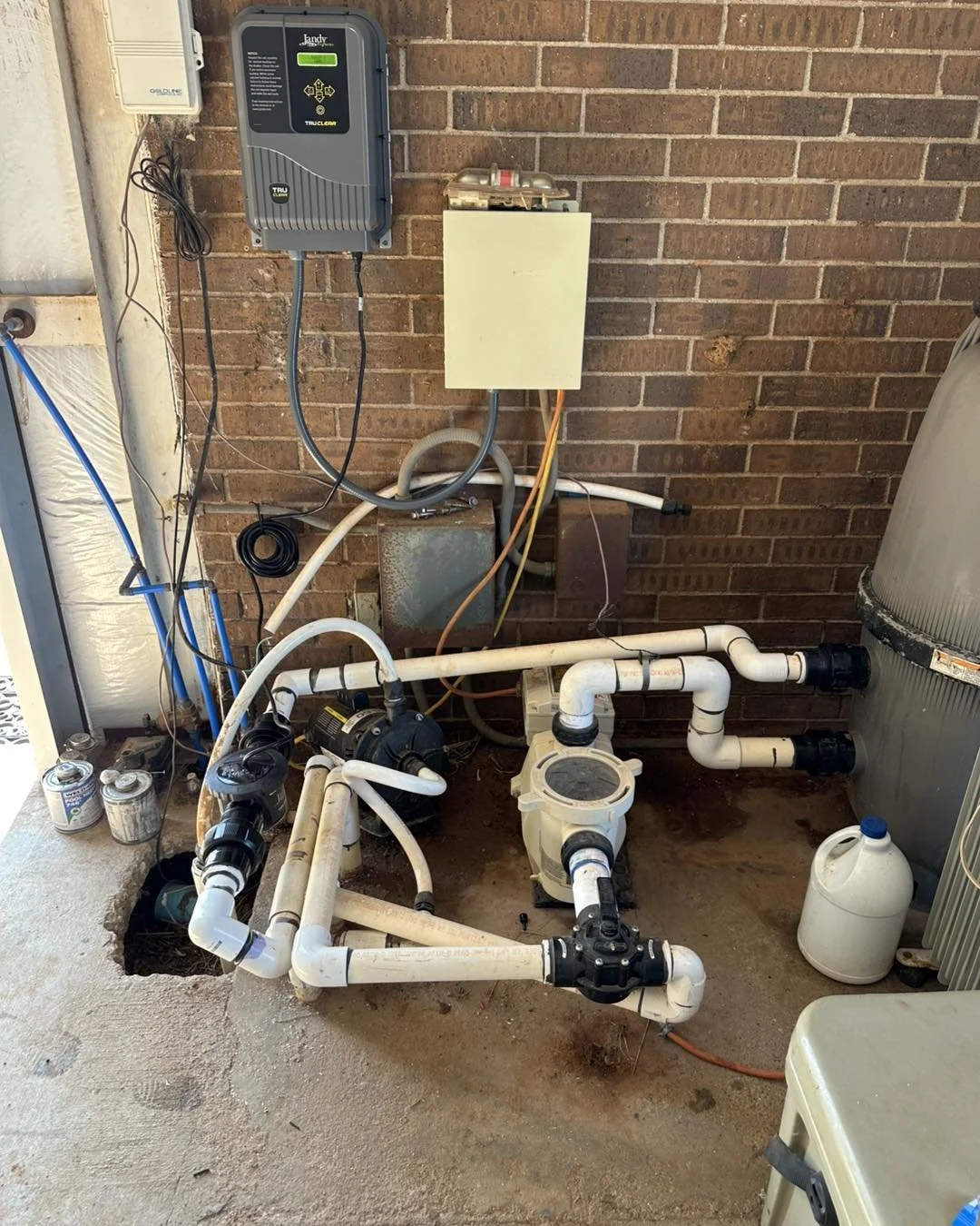 An outdoor pool pump and filtration system attached to a brick wall with various pipes, a control panel, and associated wiring.