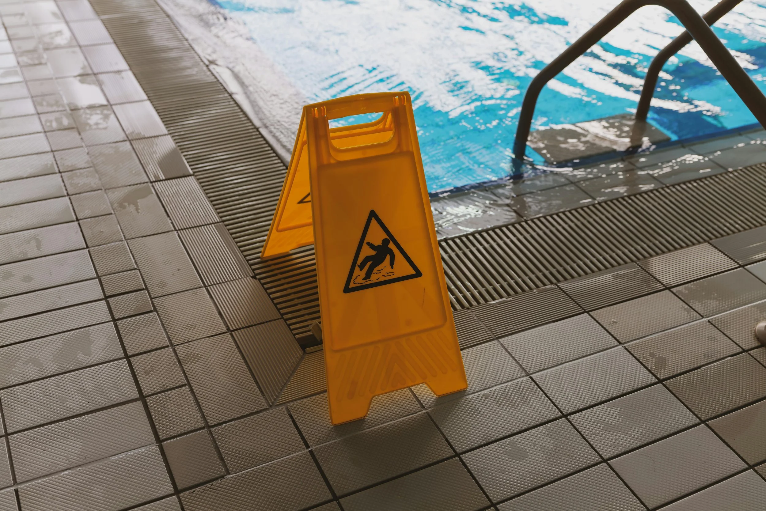 Wet tiled floor next to indoor pool with a yellow 'Caution: Slippery When Wet' sign.