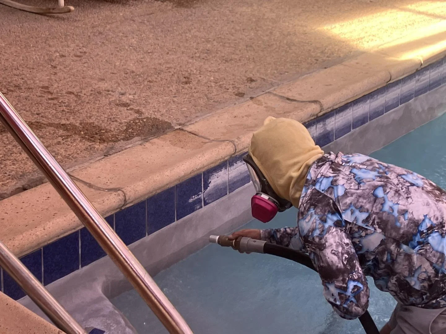 Person cleaning a pool with a high-pressure water hose, wearing a camouflage jacket, yellow hoodie, and headphones, bending over near the pool edge.
