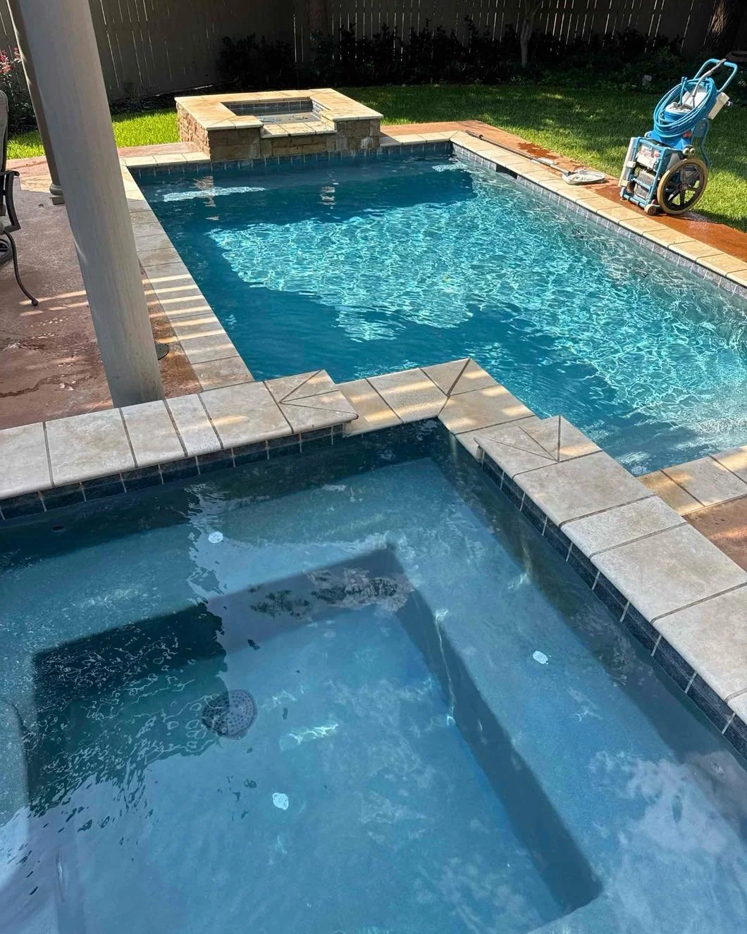 View of a backyard swimming pool with a hot tub in the foreground, concrete deck, green grass, a fenced yard, and pool cleaning equipment on the side.