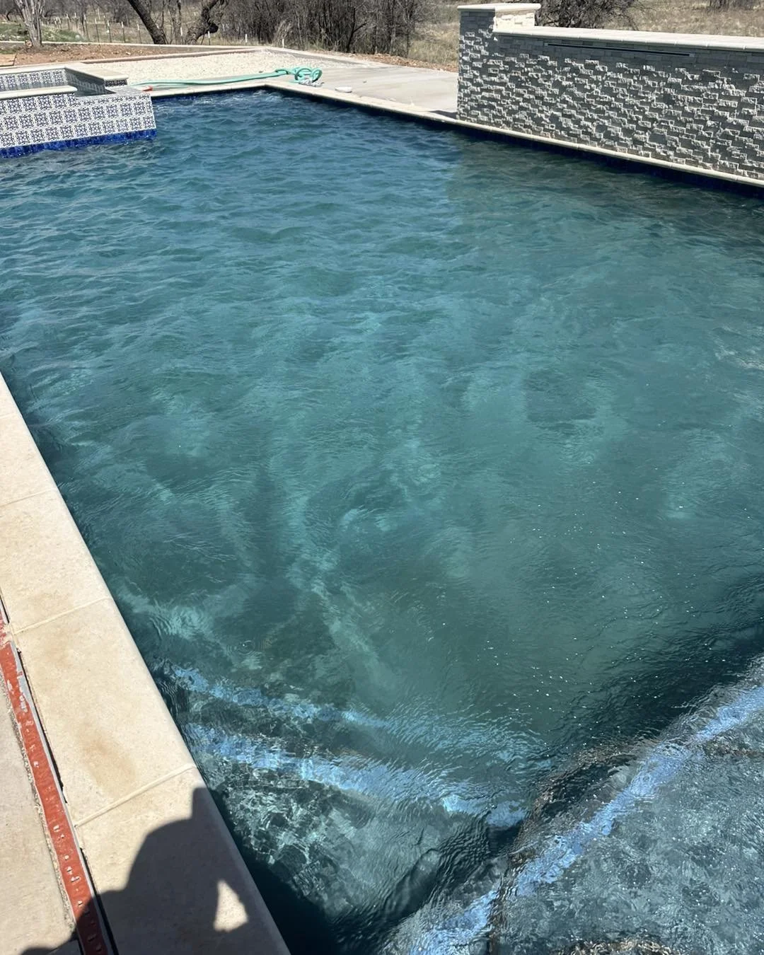 A swimming pool with clear blue water, surrounding beige tiles, and a textured grey wall on one side. There is a pool cleaner or hose on the pool deck.