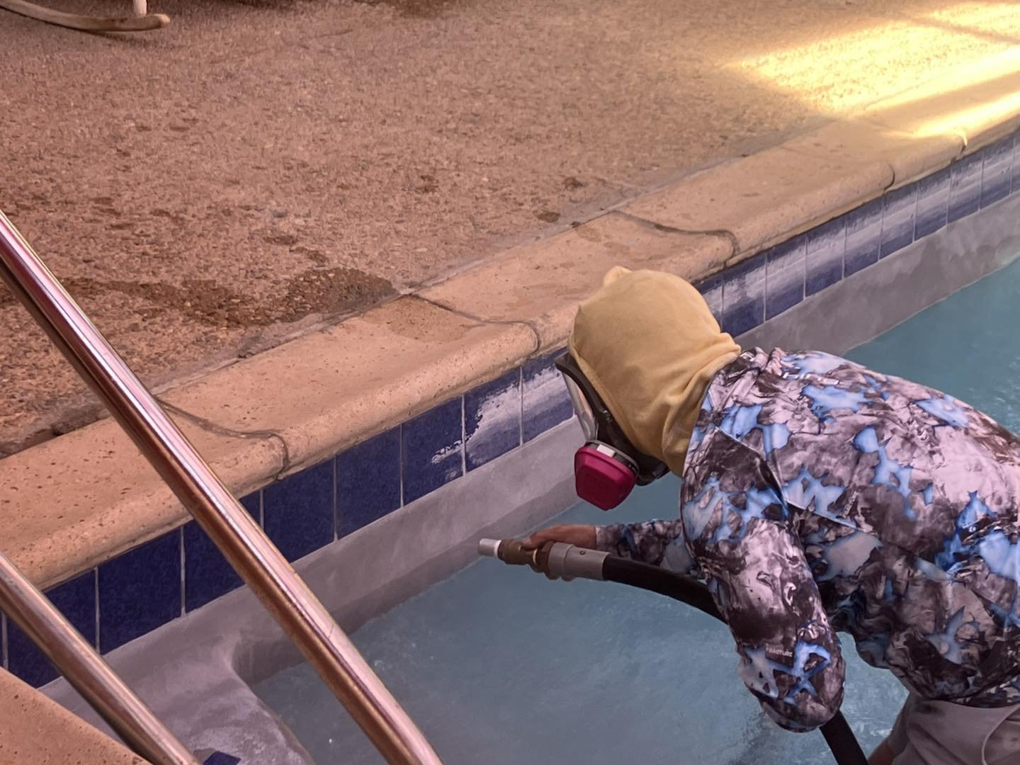 A child wearing a yellow hood, a camouflage patterned jacket, and pink ear protection is cleaning or performing maintenance on a swimming pool using a long tool.
