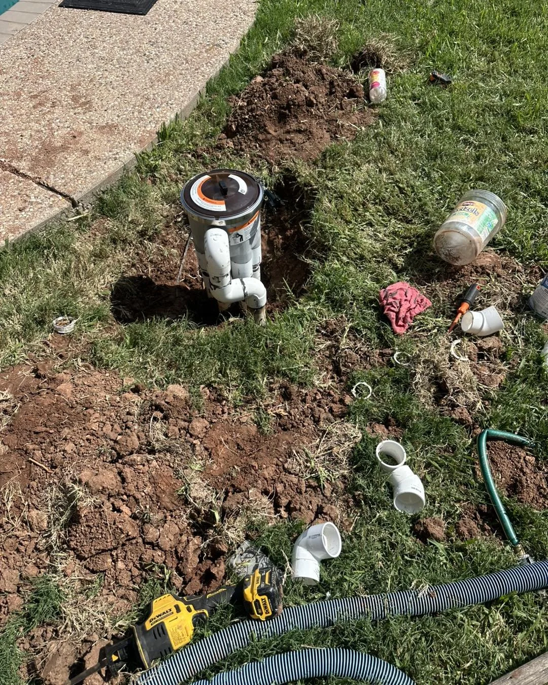 A plumbing repair in progress with PVC pipes, tools, and debris on a grassy area near a concrete sidewalk.