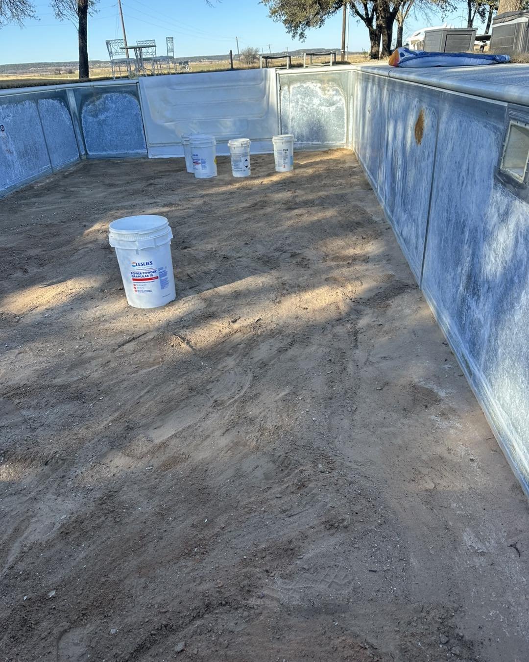 A construction site with a metal pool frame filled with dirt, surrounded by buckets of powder, and in an outdoor area with trees and open land visible in the background.