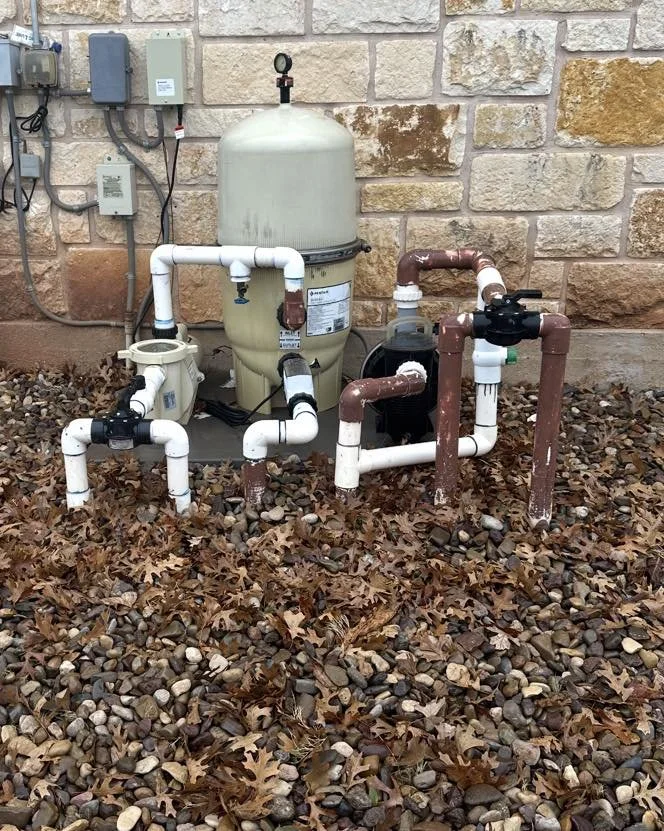 Outdoor pool filtration system with white and brown pipes on a rocky ground with fallen leaves, set against a brick wall.