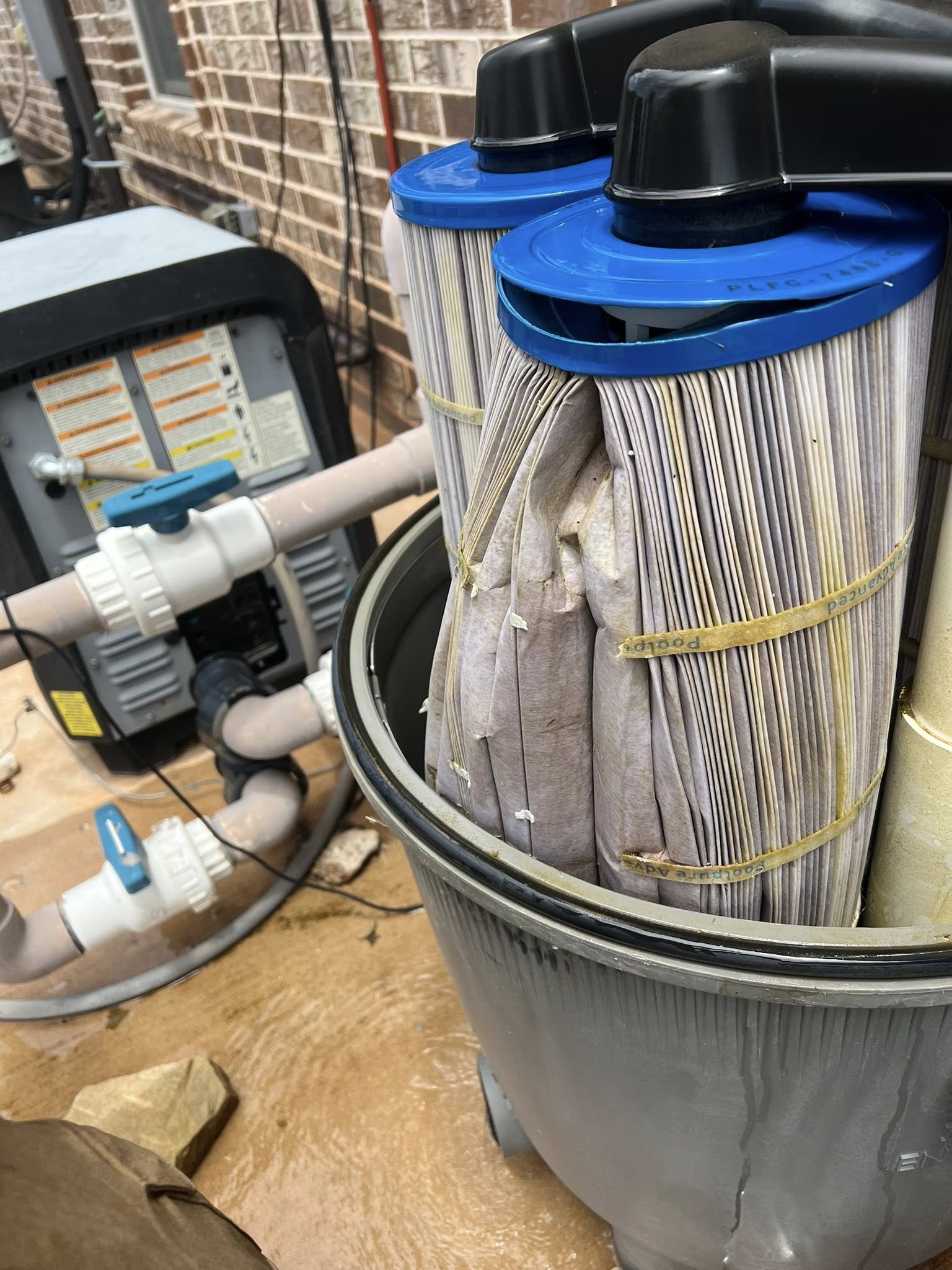 Pool filters with paper covers in a gray pool filter tank, with plumbing and a pool pump in the background.