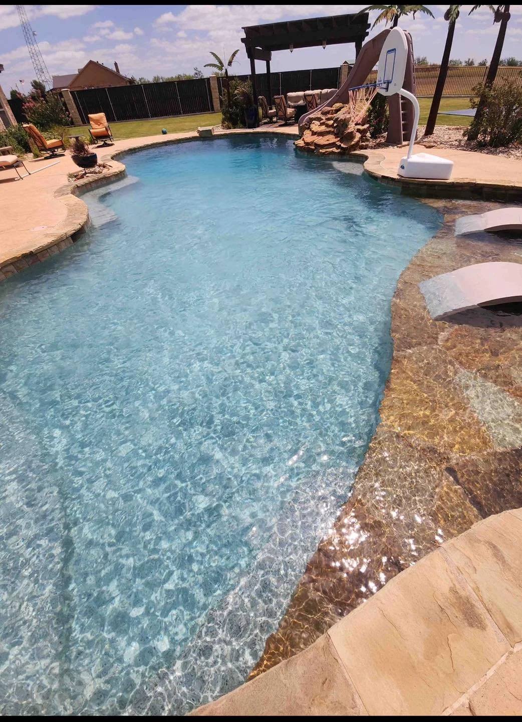 A backyard swimming pool with a slide, basketball hoop, and lounge chairs around the pool.