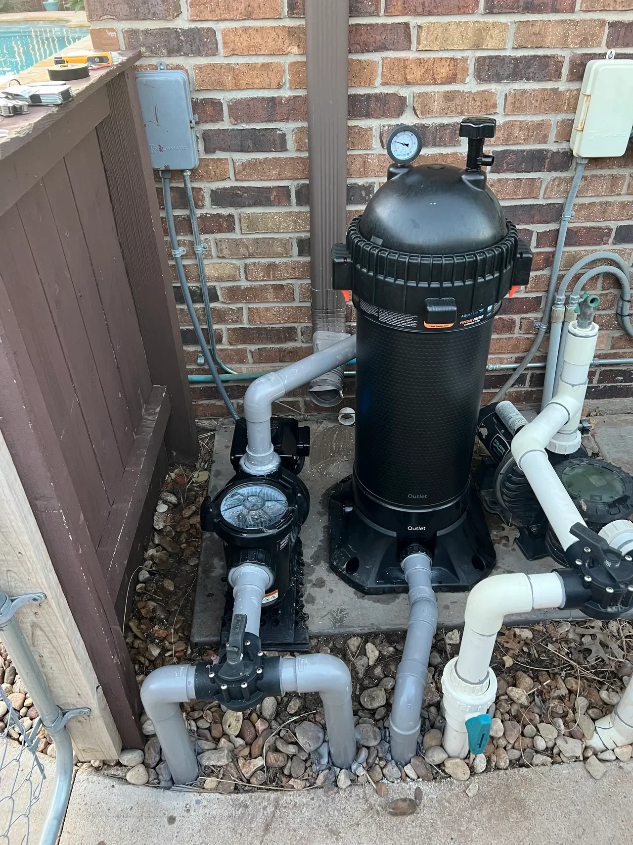 An above-ground pool filter system with a large black filter, a smaller pump, and various pipes and gauges installed outside on a concrete slab near a brick wall.